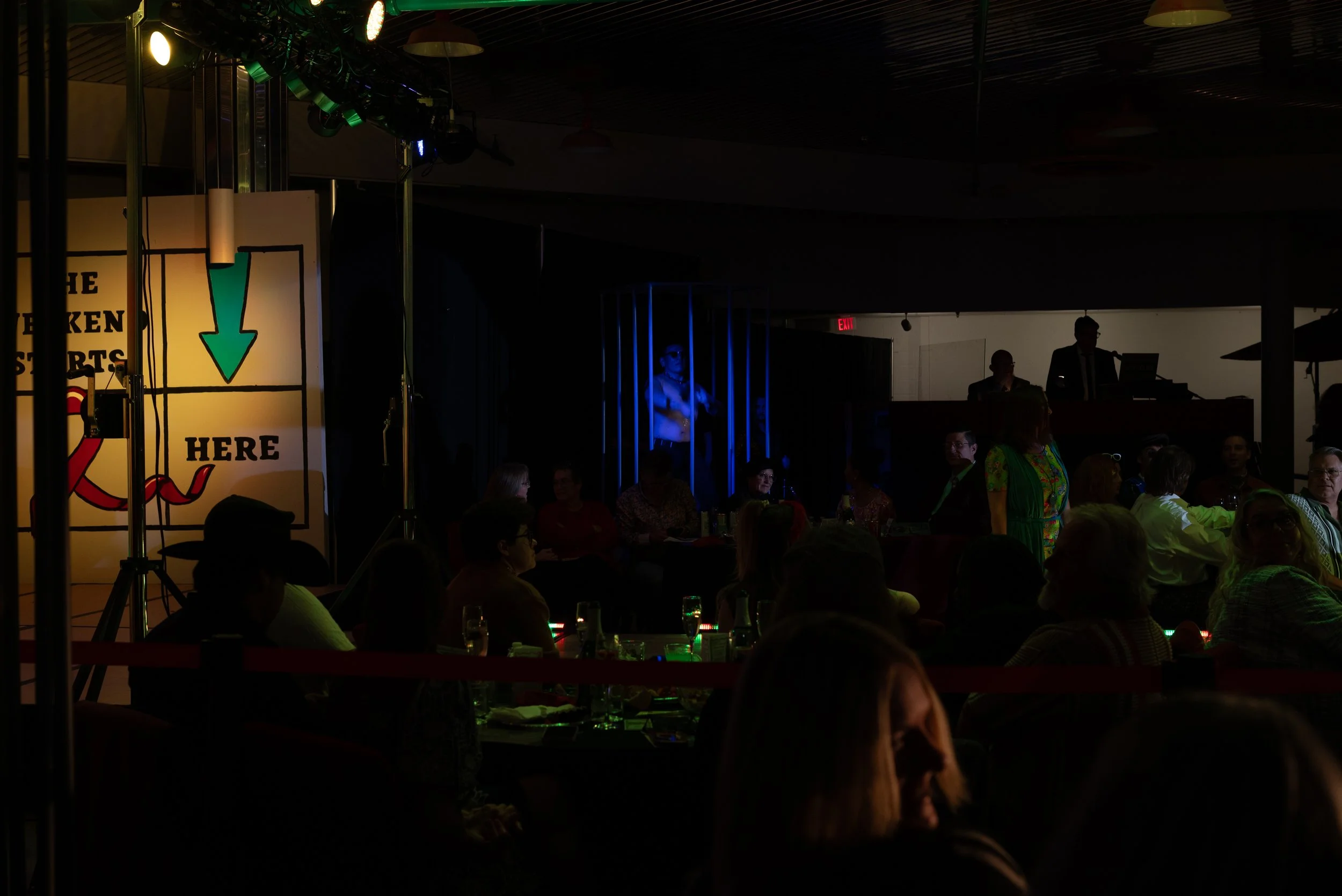 A crowd seated at tables in a dark indoor venue, with a small stage on the left featuring a sign that says 'HERE' and an arrow pointing downward. The stage area is poorly lit, with some people standing and a person in sunglasses performing. There are