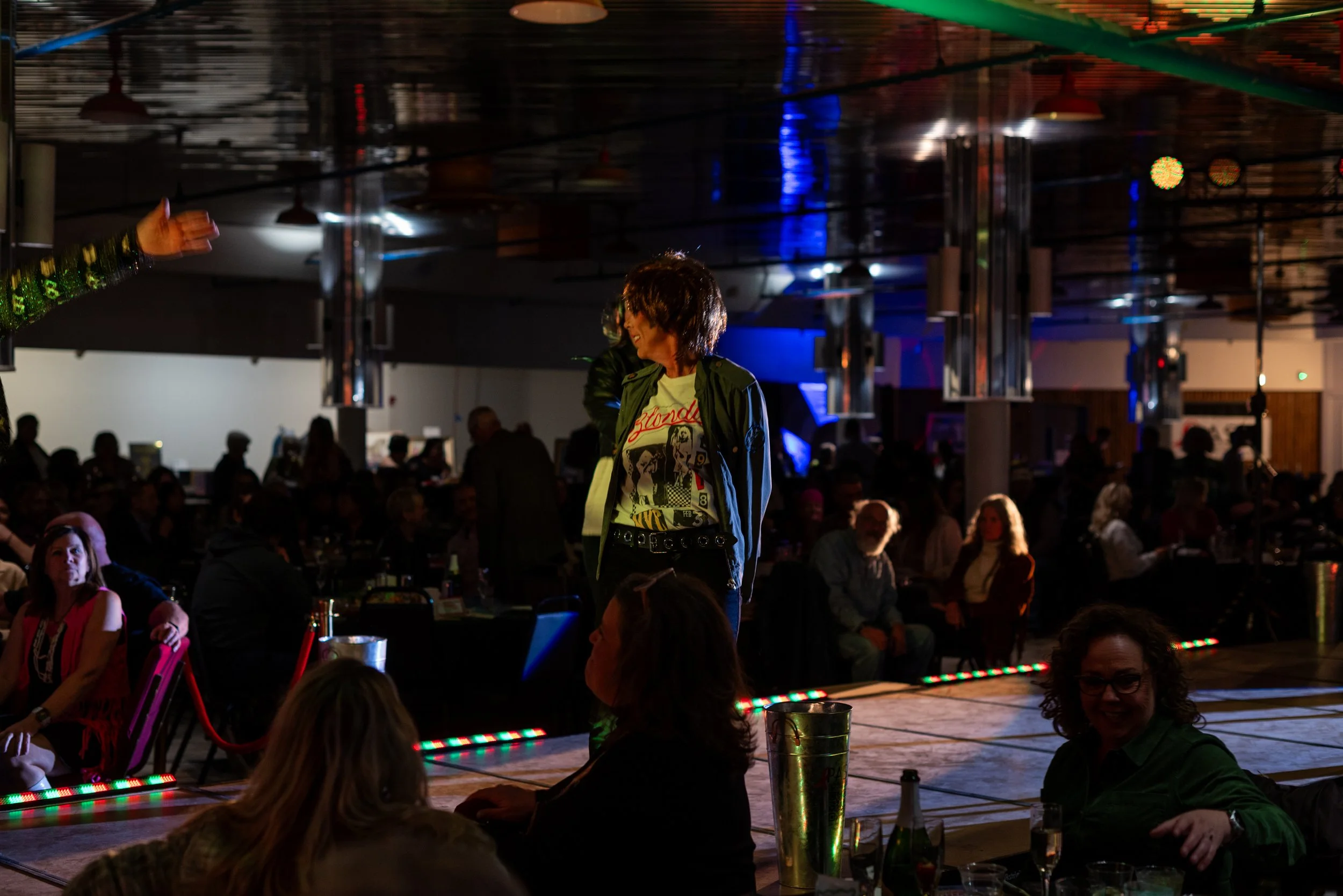 A woman with curly hair wearing a graphic t-shirt, a green jacket, and black pants stands on a stage in a dimly lit event space. The background shows an audience sitting at tables, some with drinks, and the ceiling has reflective surfaces with colorf