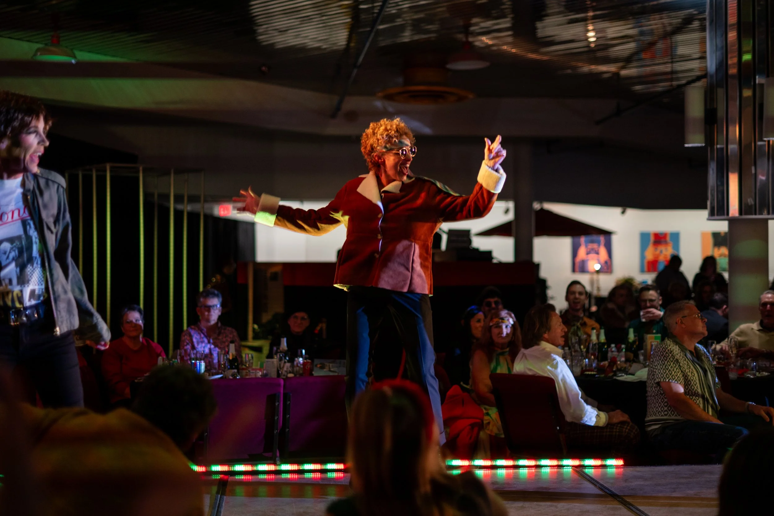 A woman with gray curly hair, glasses, and a red and white jacket is dancing on stage at an event with an audience watching and smiling, seated at tables.
