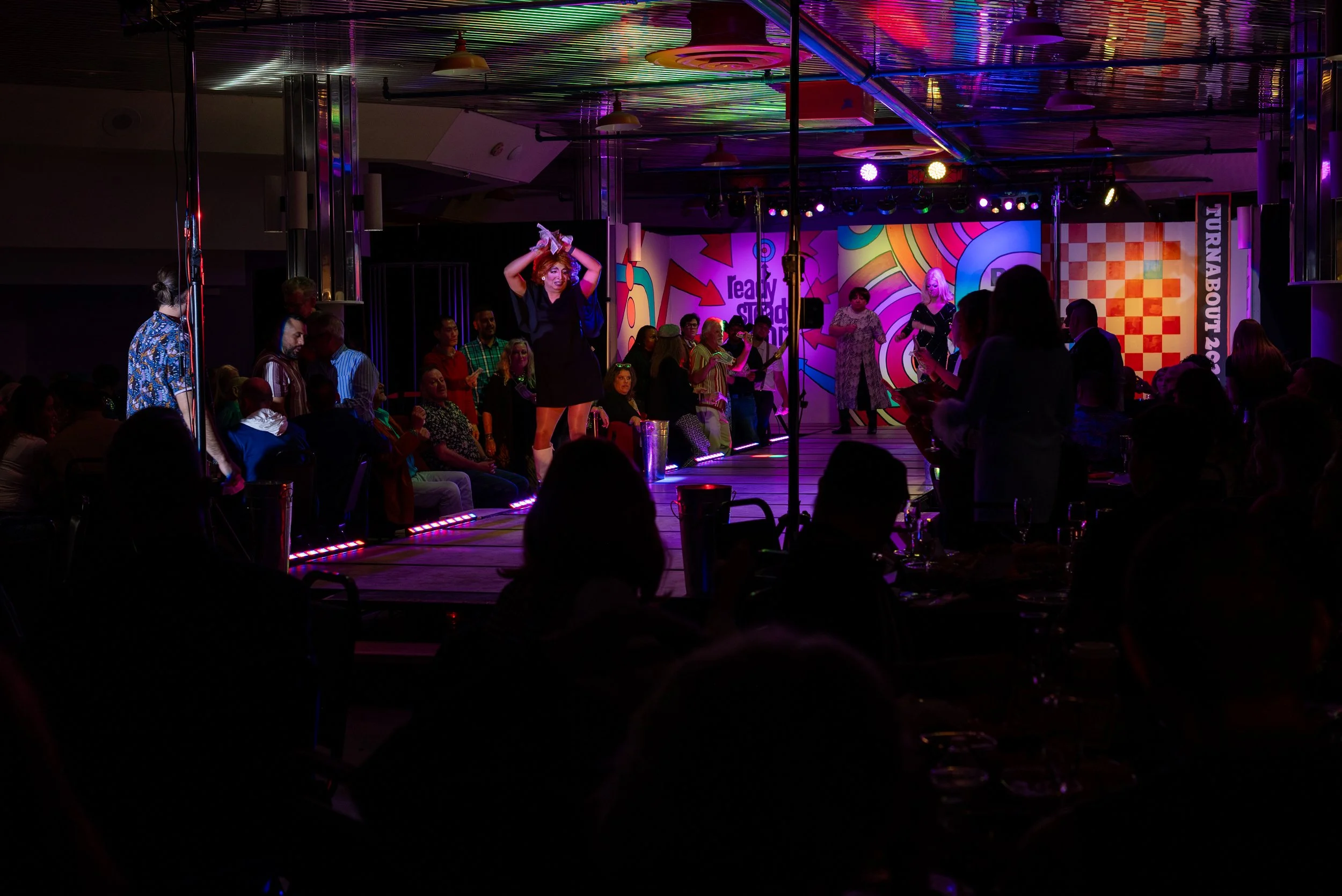 Stage with performers and audience at a lively drag show, colorful background, and vibrant lighting.