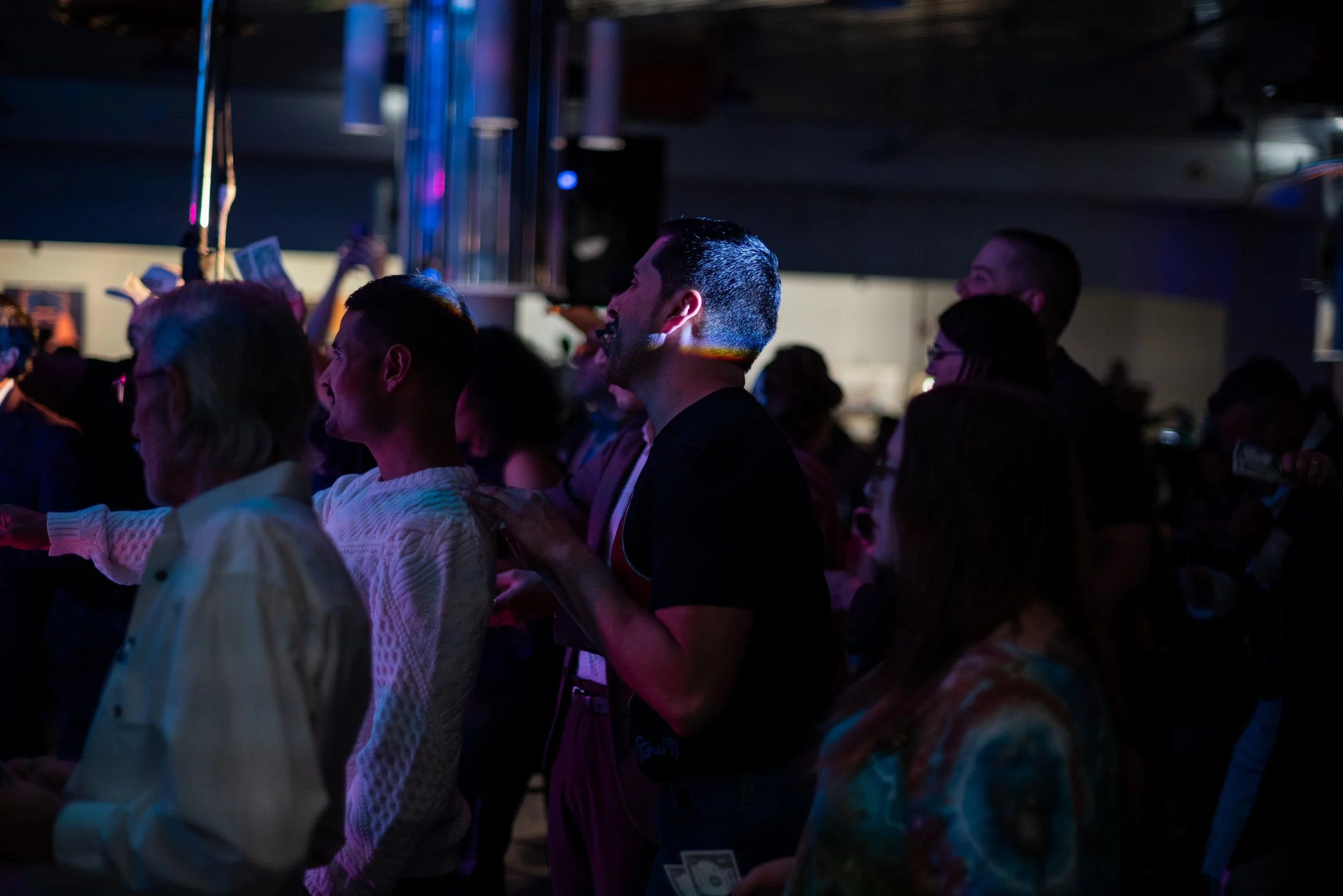 Group of people at a lively indoor concert or event, standing close together, enjoying music under dim lighting with colorful stage lights in the background.