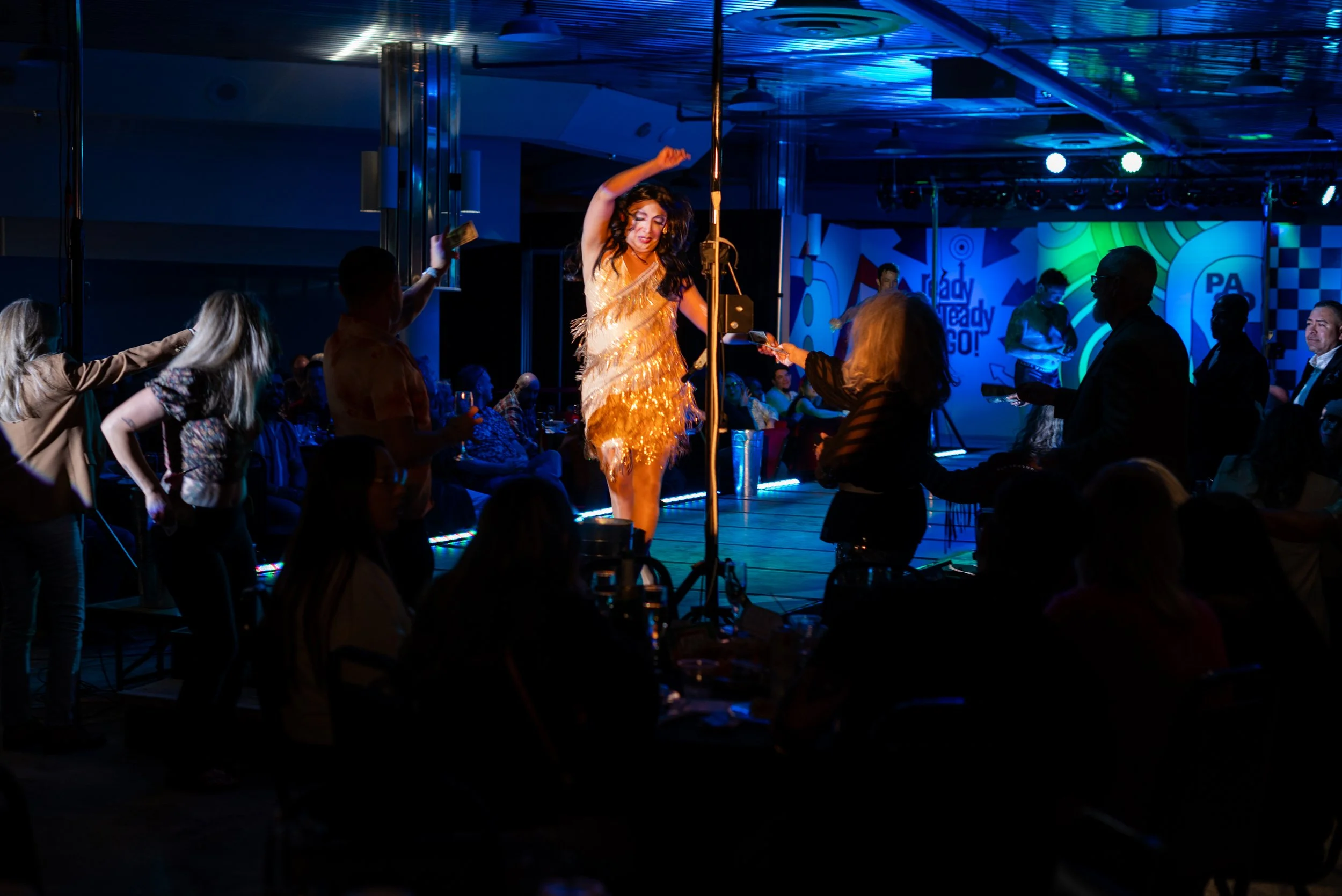 A woman in a gold, feathered dress singing or performing on a stage at a lively party or event, with people dancing and enjoying themselves around her.