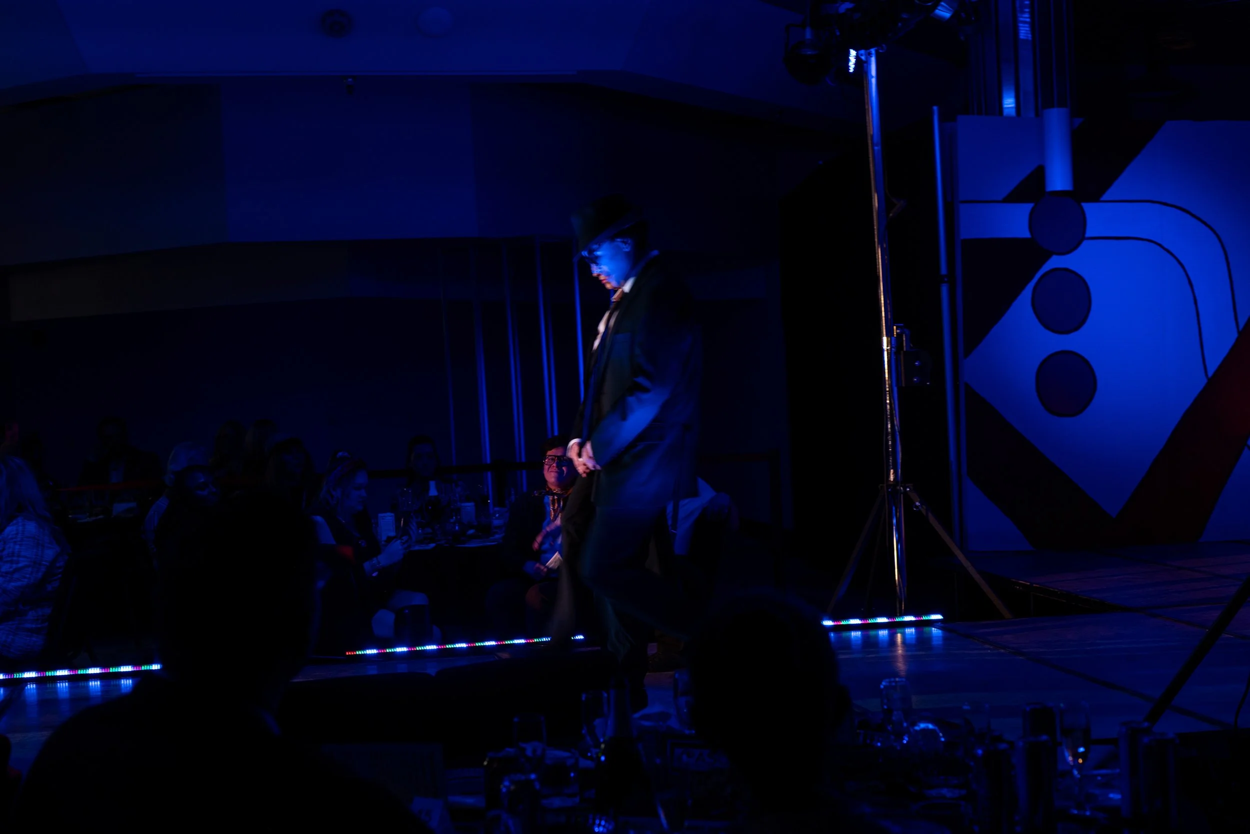 A man dressed in a black suit and hat stands on a stage with a lit-up background featuring dice graphic, while an audience sits at tables watching a performance or event in a dimly lit room.
