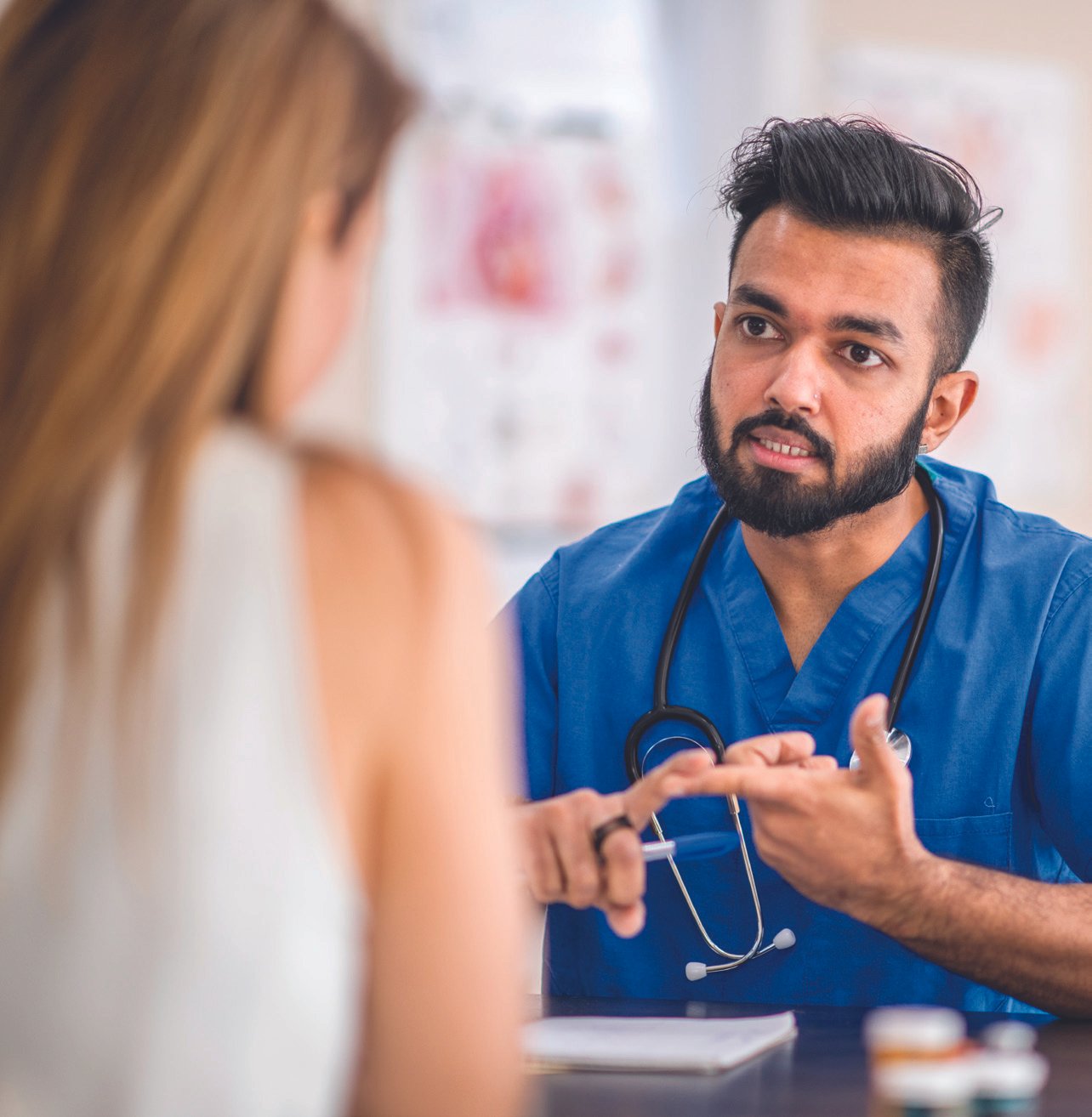 A male healthcare professional wearing blue scrubs and a stethoscope discusses with a female patient in a medical setting.