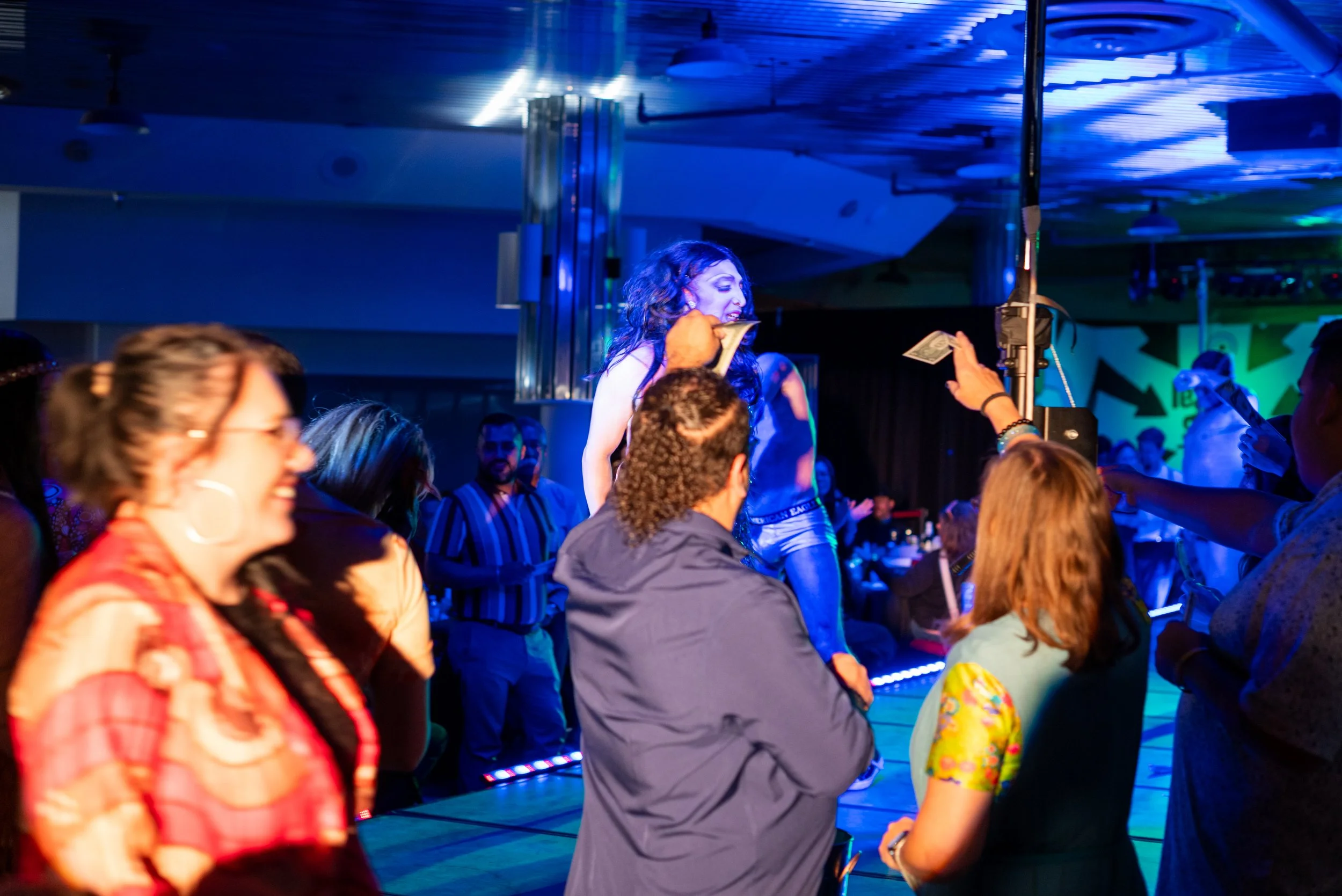 A lively indoor event with people dancing and socializing under colorful lighting, with one woman singing on a stage.