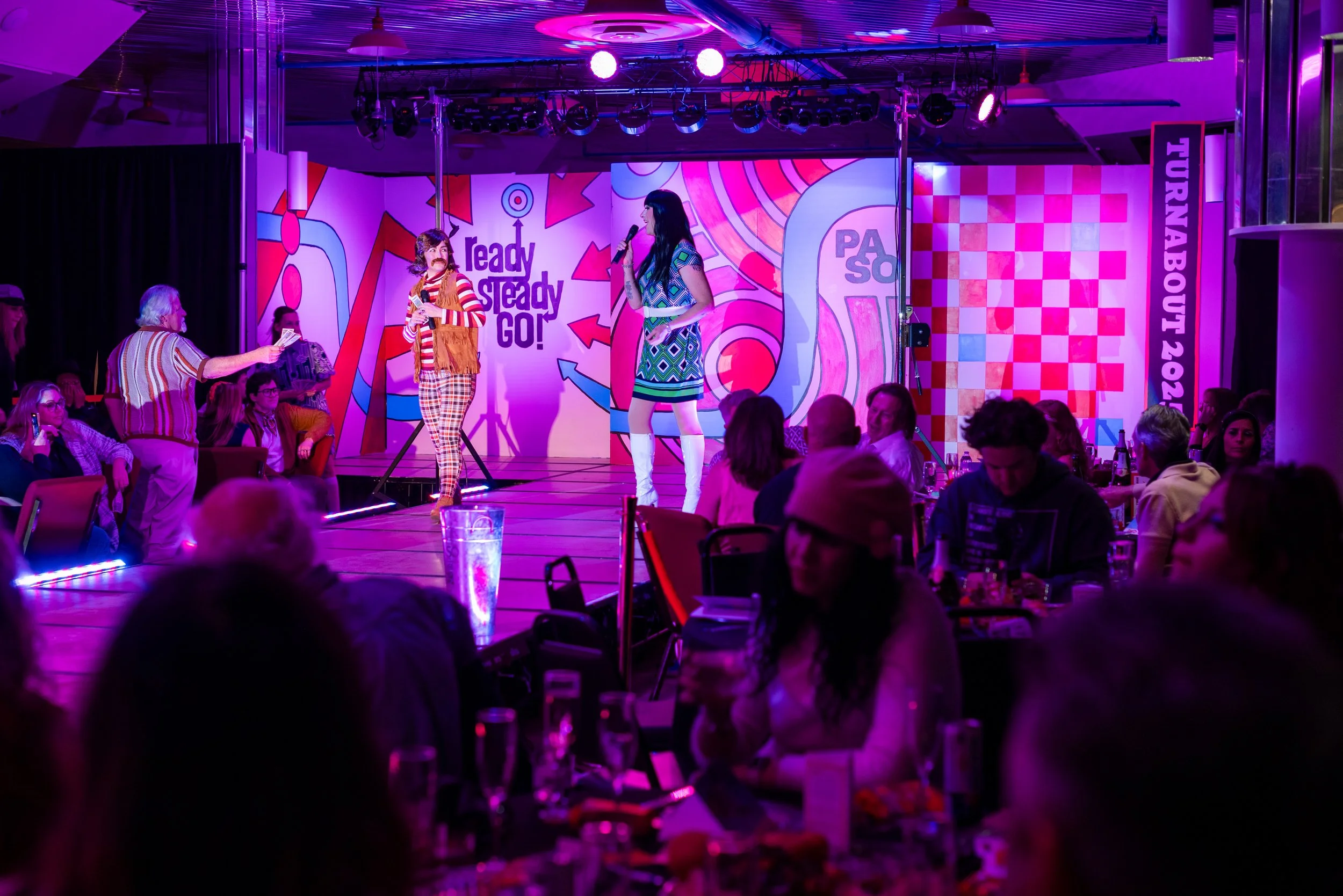 People on stage performing comedy or acting in front of an audience in a dimly lit, colorful venue with purple and pink lighting.