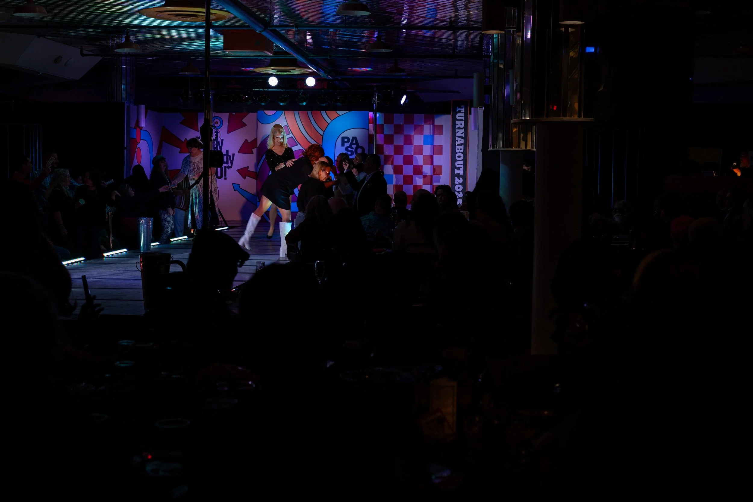 Stage with performers and audience in a dark room, with colorful banners reading 'Turnabout 2023' and 'PASA' in the background.