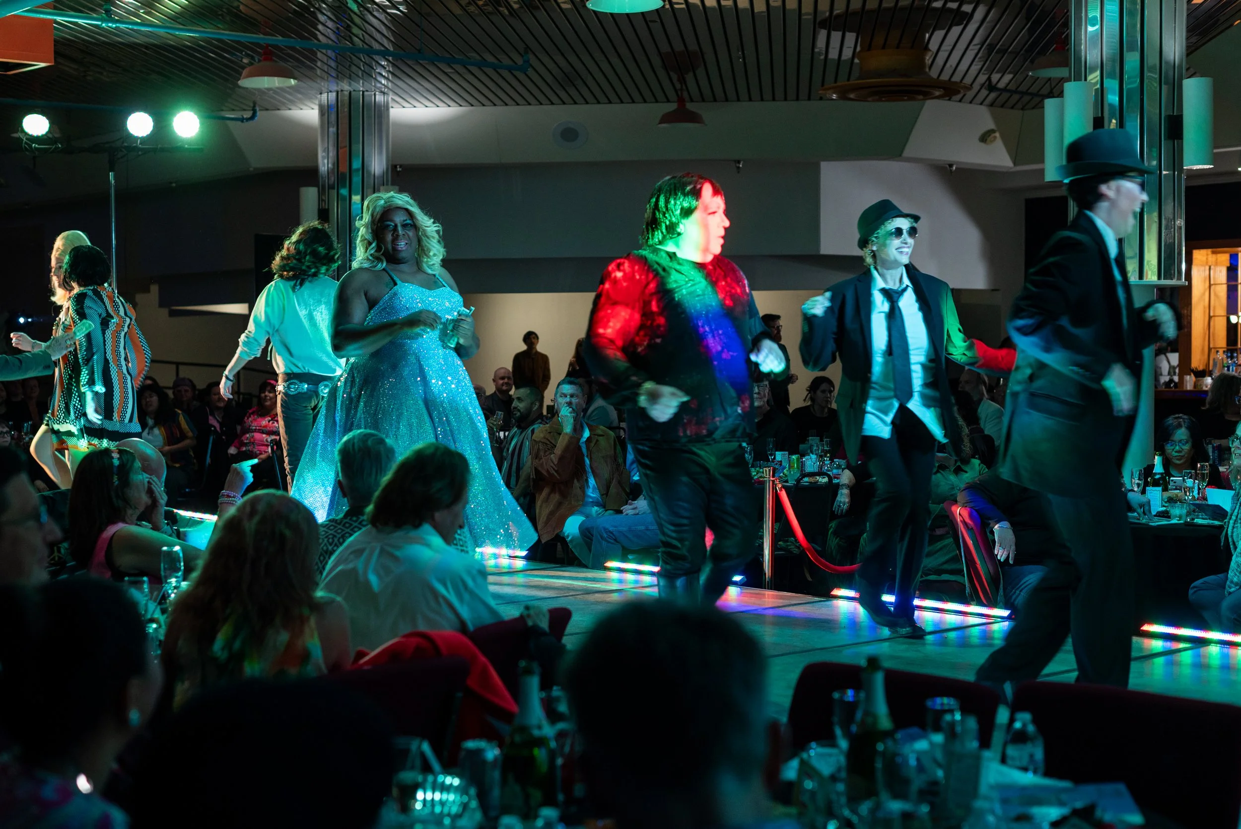 People dancing on a stage at a party or event, with some dressed in colorful and shiny clothing, while others are dressed in suits and hats, with an audience watching in the background.