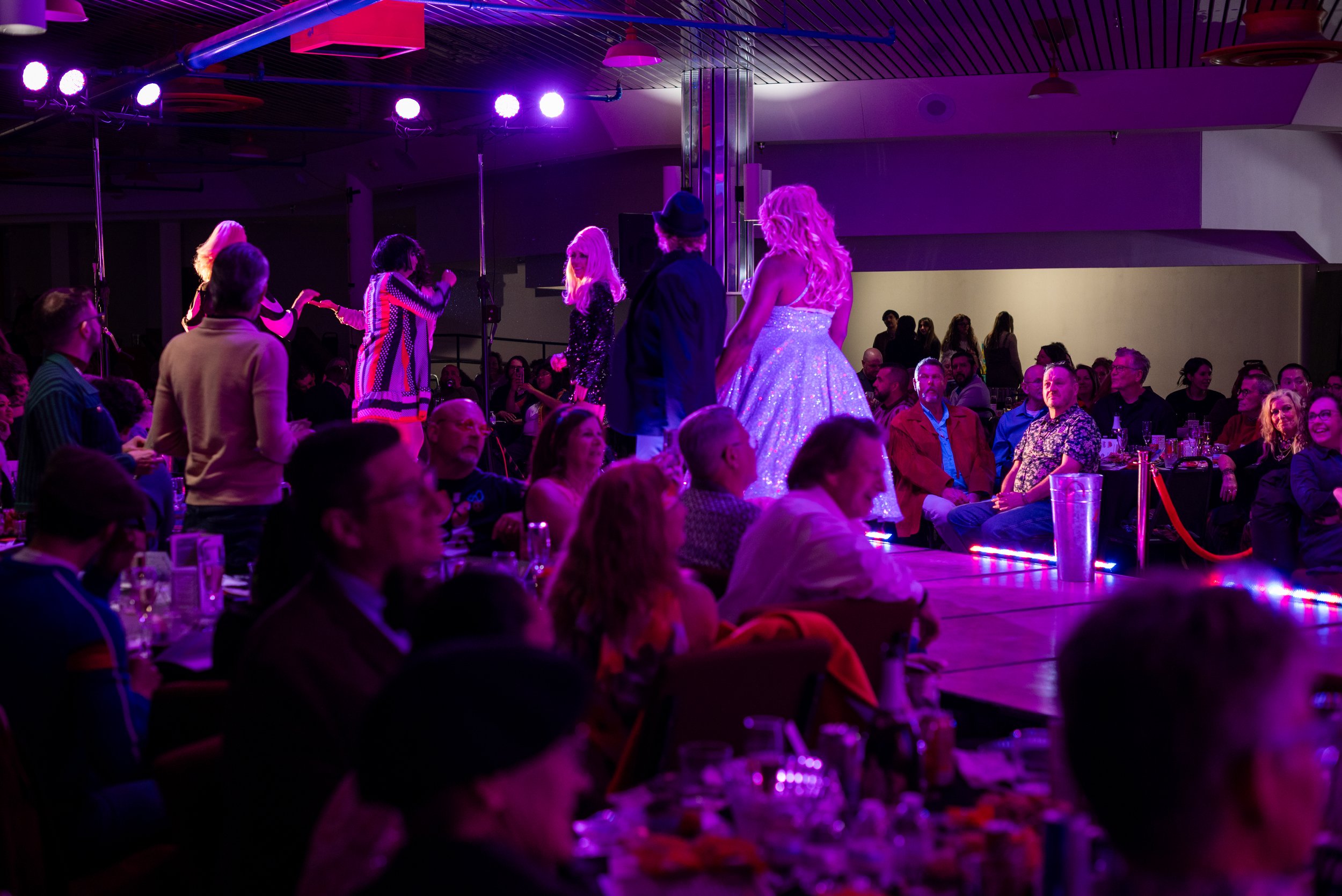 Audience watching a fashion show with models on runway, illuminated by purple and pink stage lights, in a dark indoor venue.
