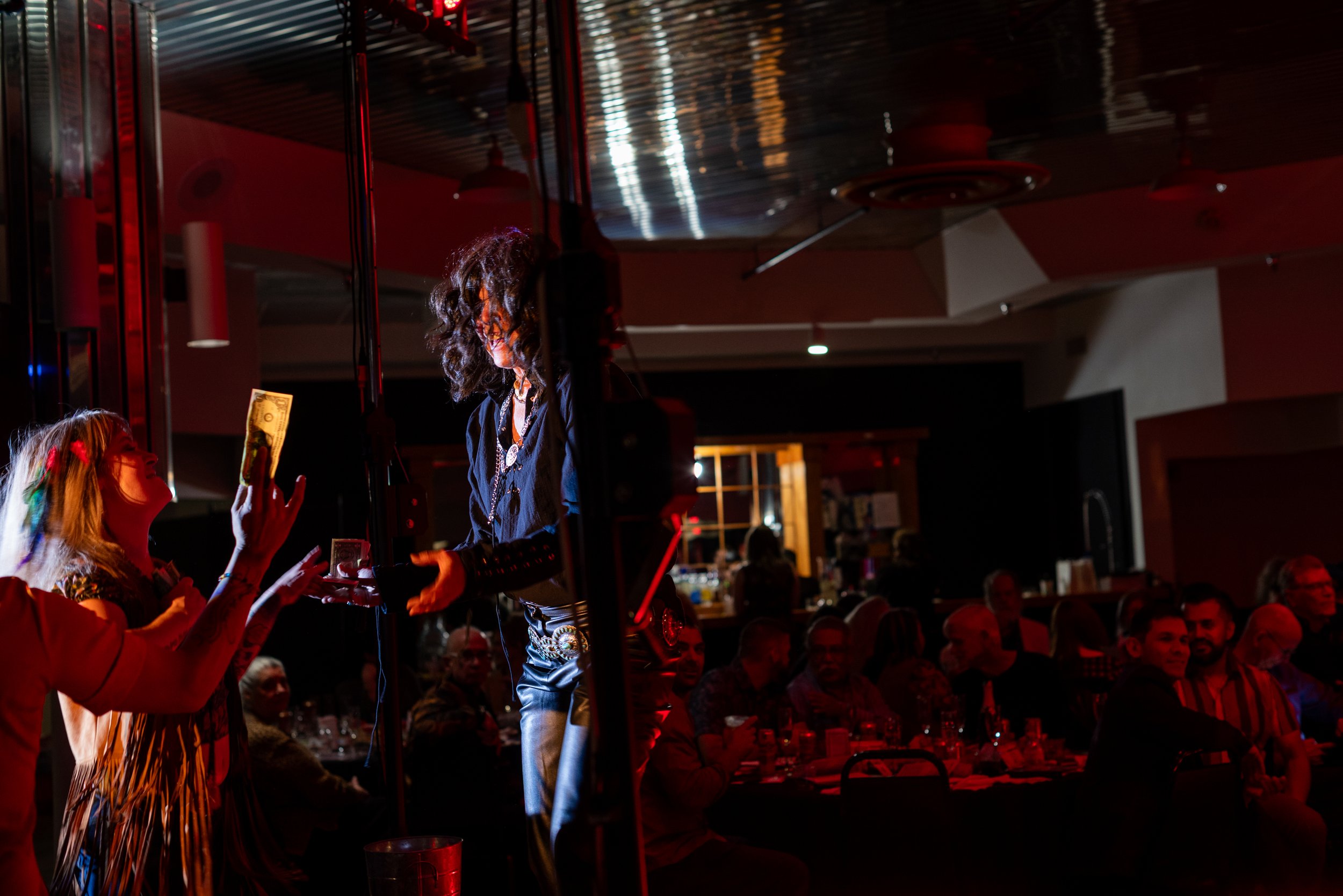 A woman on stage receiving money from an audience member during a performance in a dimly lit venue with audience sitting at tables.