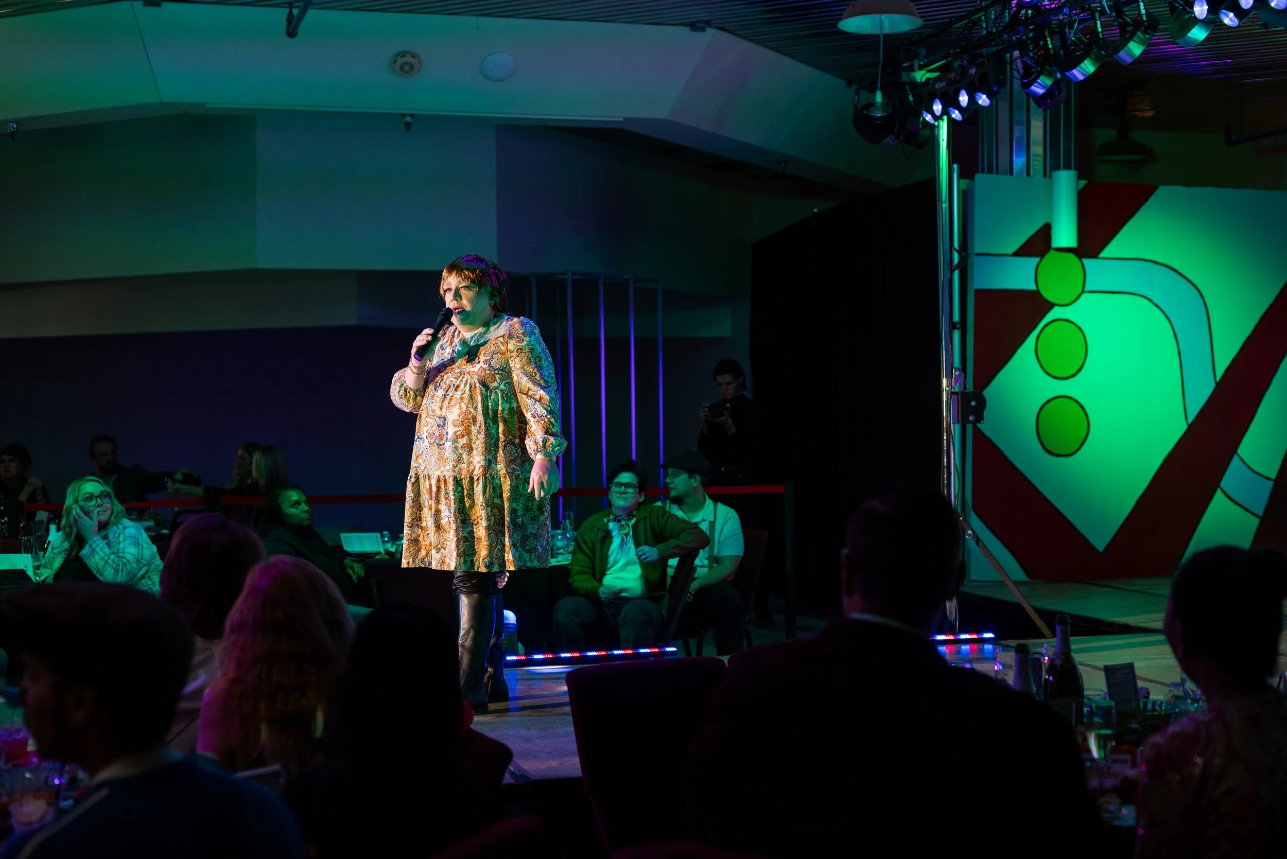 A woman performs stand-up comedy on stage, holding a microphone, with an audience seated around tables in a dimly lit venue.