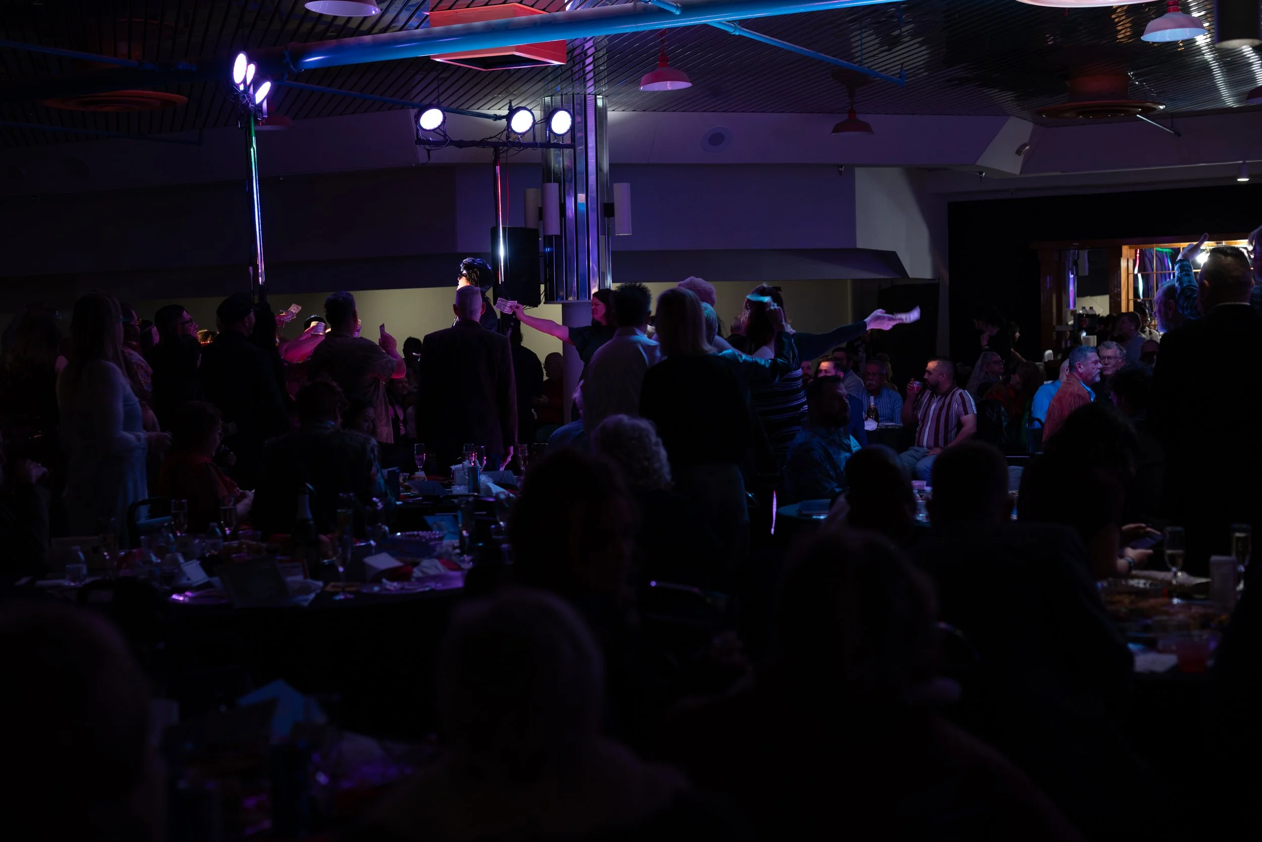 A large, dark indoor event space filled with people sitting and standing. There is a stage with performers or speakers and bright stage lighting. The audience is engaged, some watching the stage, others talking or using their phones.