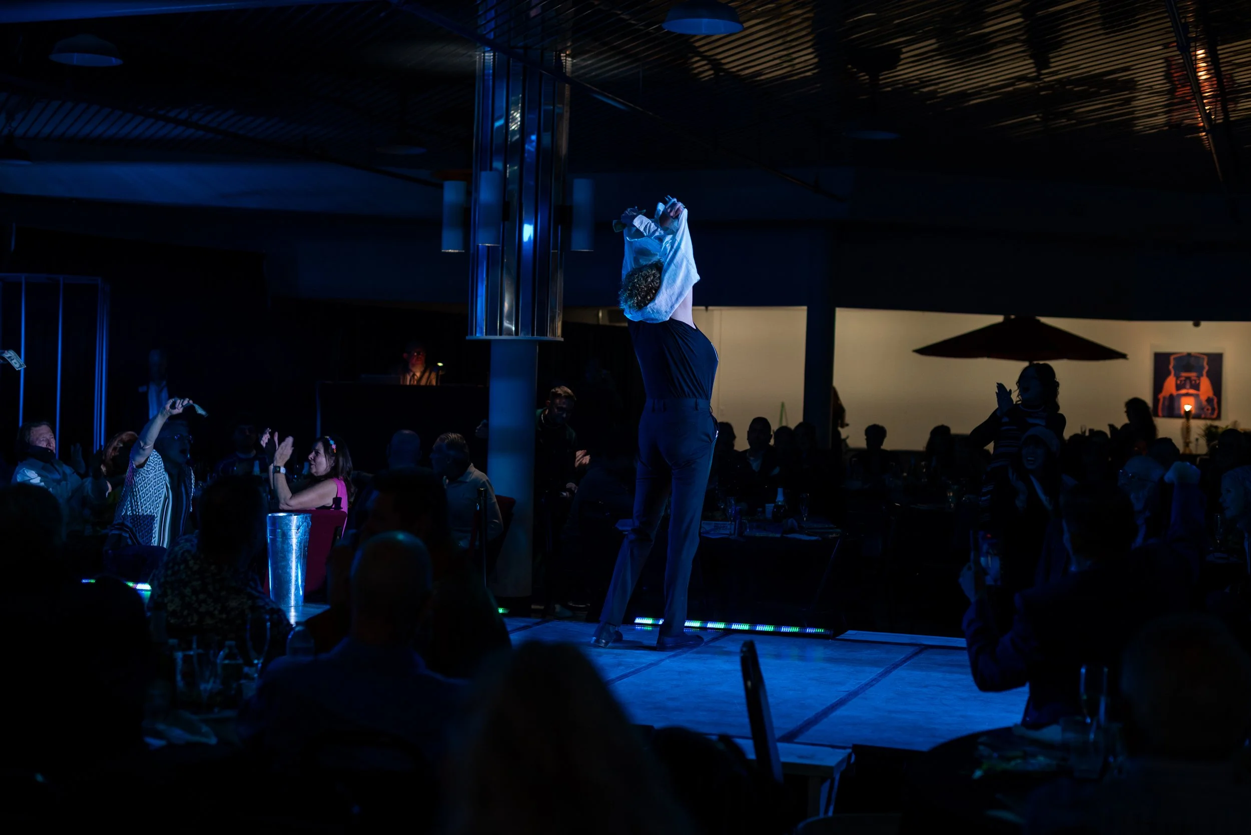 Person performing a dance move on a stage in front of an audience in a dimly lit room.