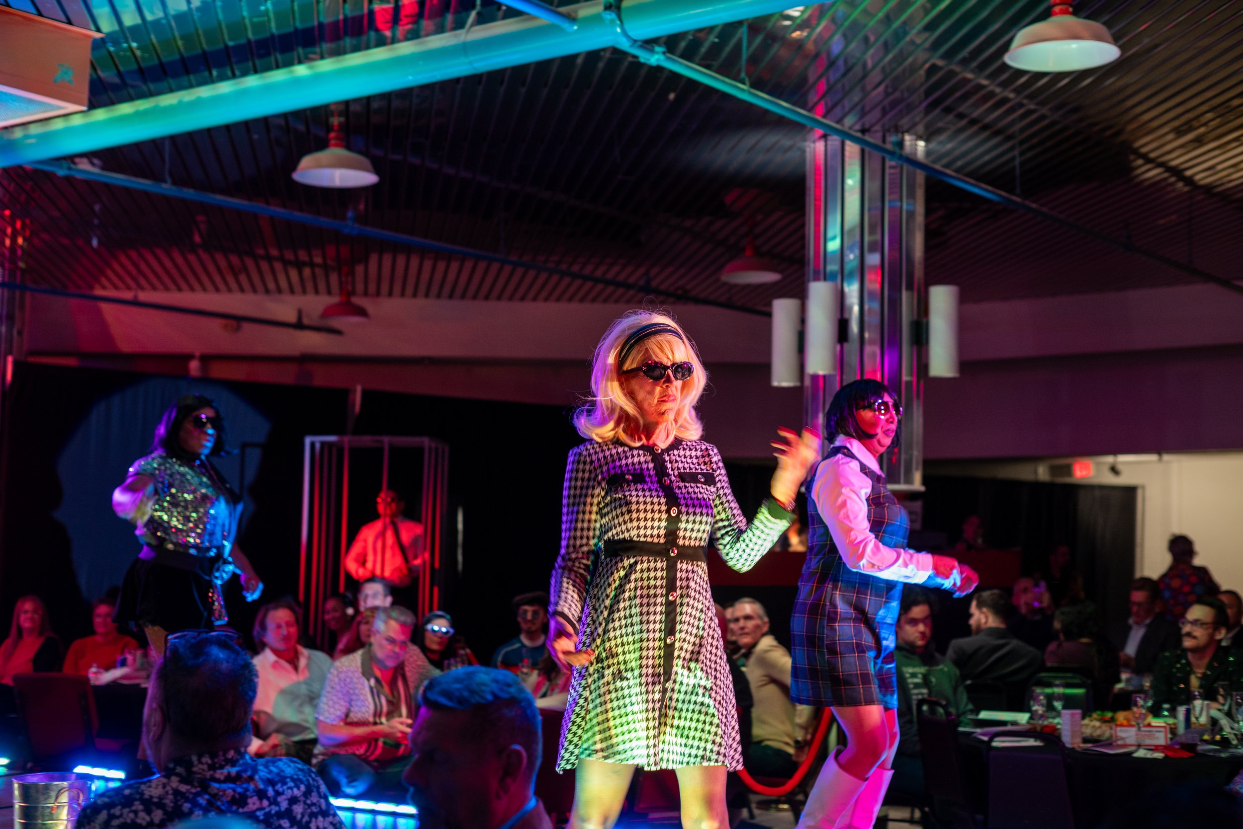 People dancing on stage at a nightclub with colorful lighting, wearing retro outfits and sunglasses, with an audience watching.