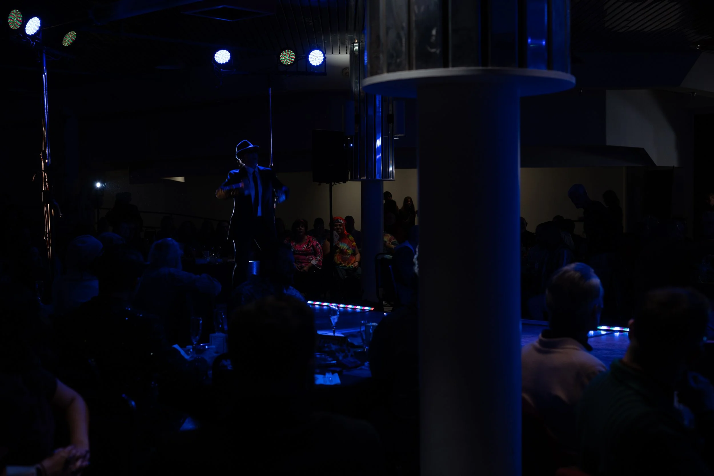 A performer on stage wearing a hat, suit, and tie, illuminated in blue lighting, with an audience seated in the dark around the stage, and colorful lights overhead.