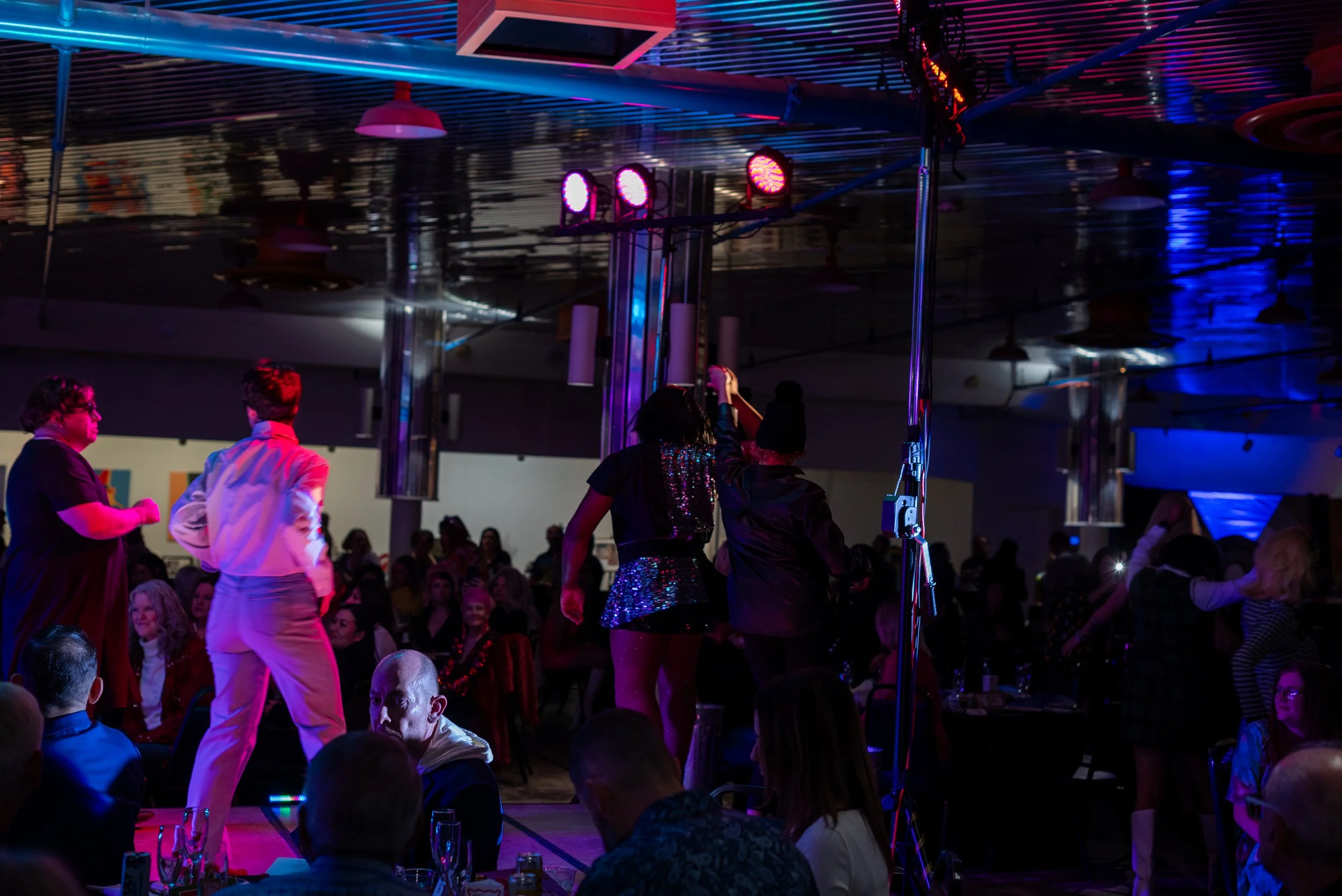 People dancing and socializing at a lively indoor event with colorful lighting and a stage.