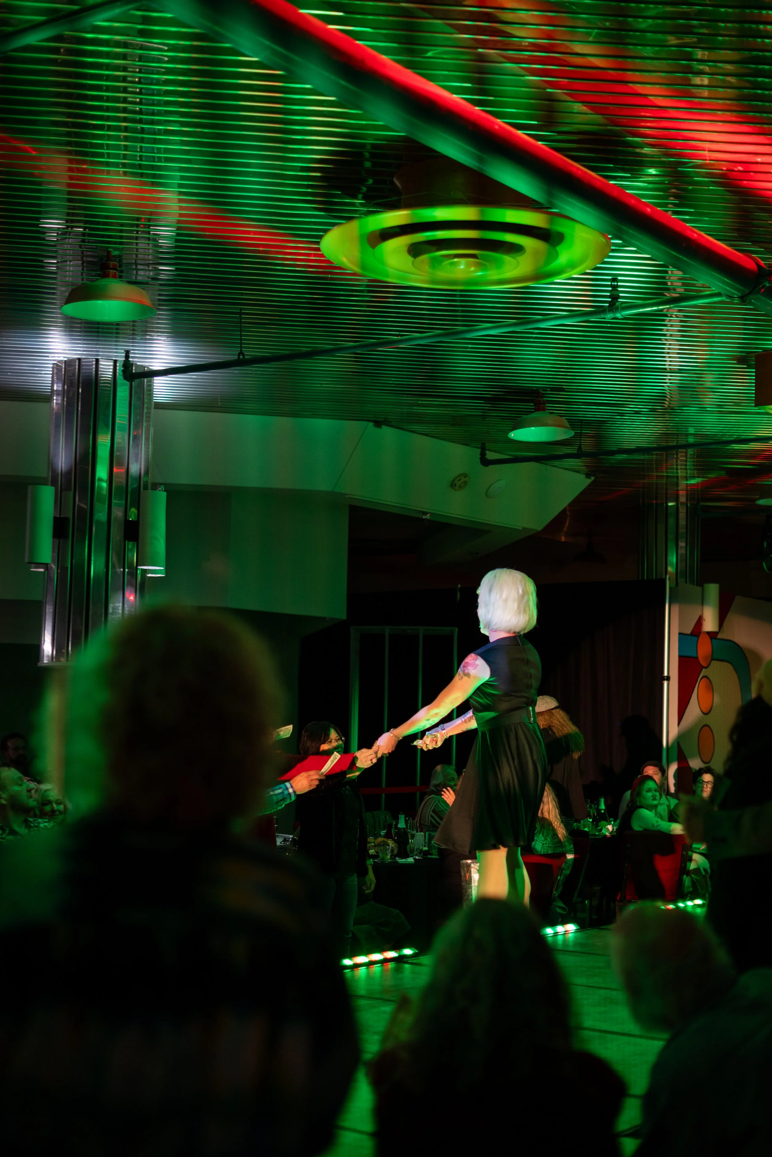 A woman with white hair in a black dress shaking hands with a person in a red shirt at a stage in a dimly lit venue with green and red lighting.