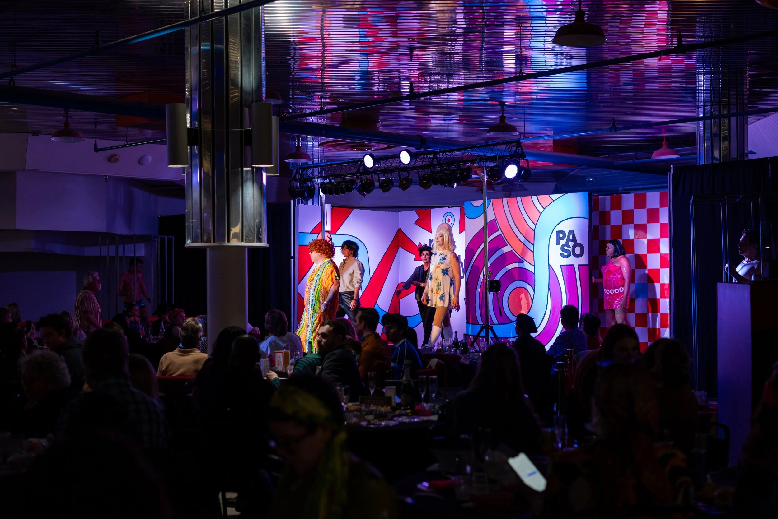 A live stage performance with five performers, colorful backgrounds, and an audience seated at tables in a dimly lit venue.