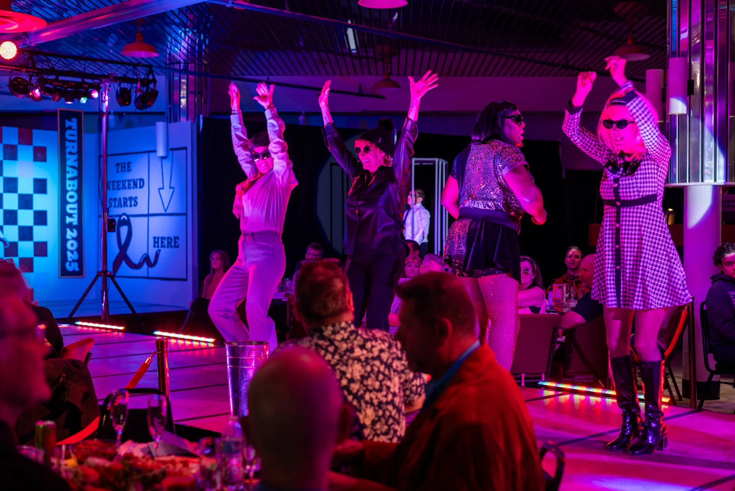 People dancing on stage at a nightclub or event, with an audience watching. The stage is lit with pink and purple lights, and people are wearing glamorous outfits and sunglasses.
