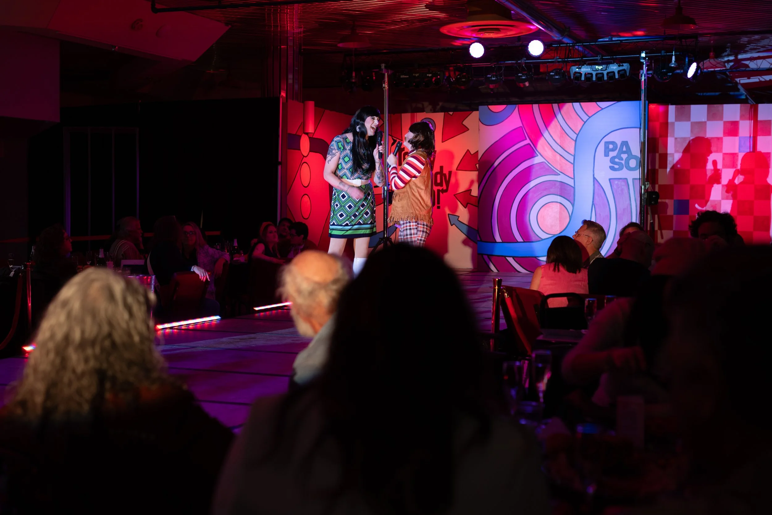 Two women are on stage performing comedy or singing at an event with a colorful, graffiti-style backdrop and audience members seated at tables watching.