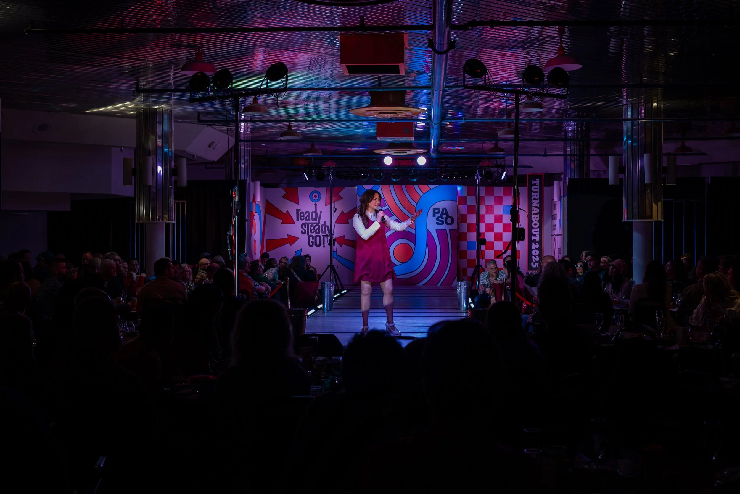 A woman performing on a stage with a microphone in a dimly lit venue, with an audience seated around. Colorful graphics and text are visible on the backdrop behind her.