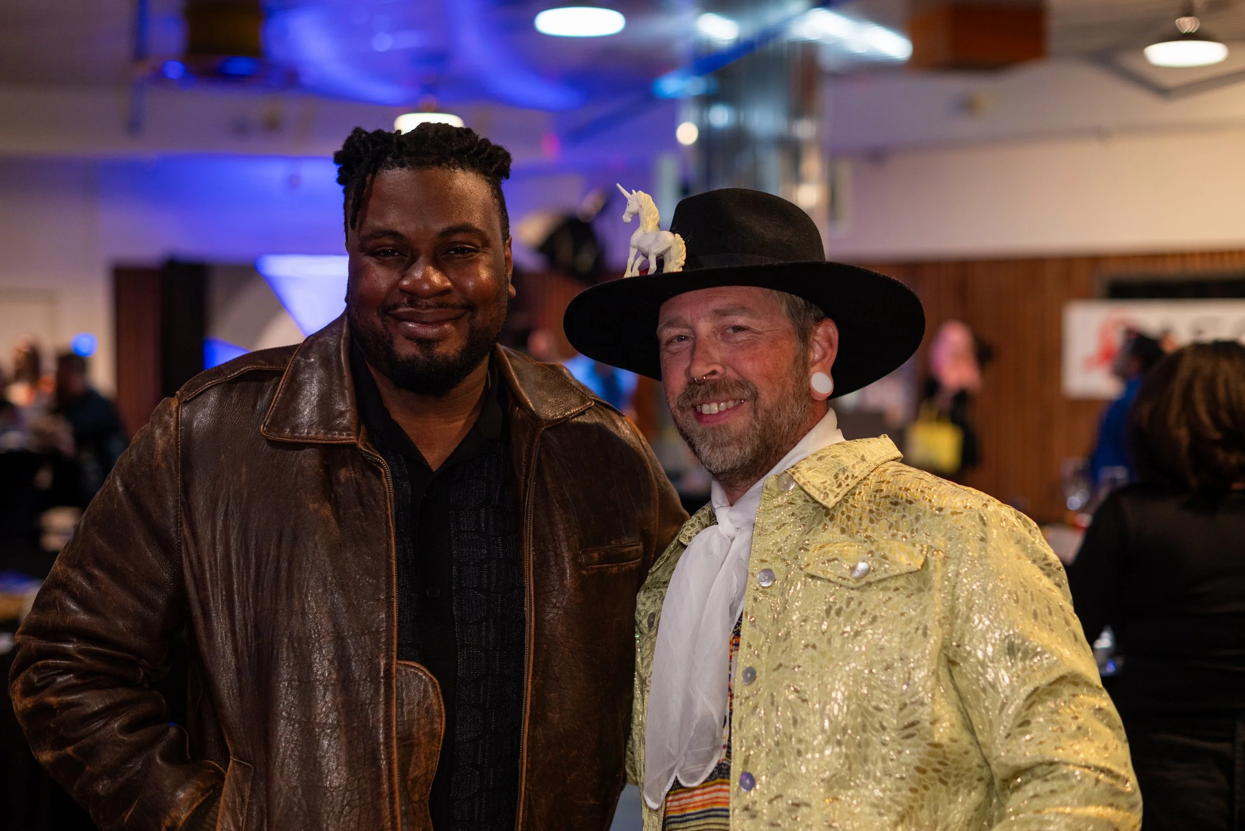 Two men smiling at a social event. The man on the left has dark skin, short curly black hair, and is wearing a brown leather jacket. The man on the right has light skin, a beard, and is dressed in a yellow ornate jacket, a white neck piece, and a lar