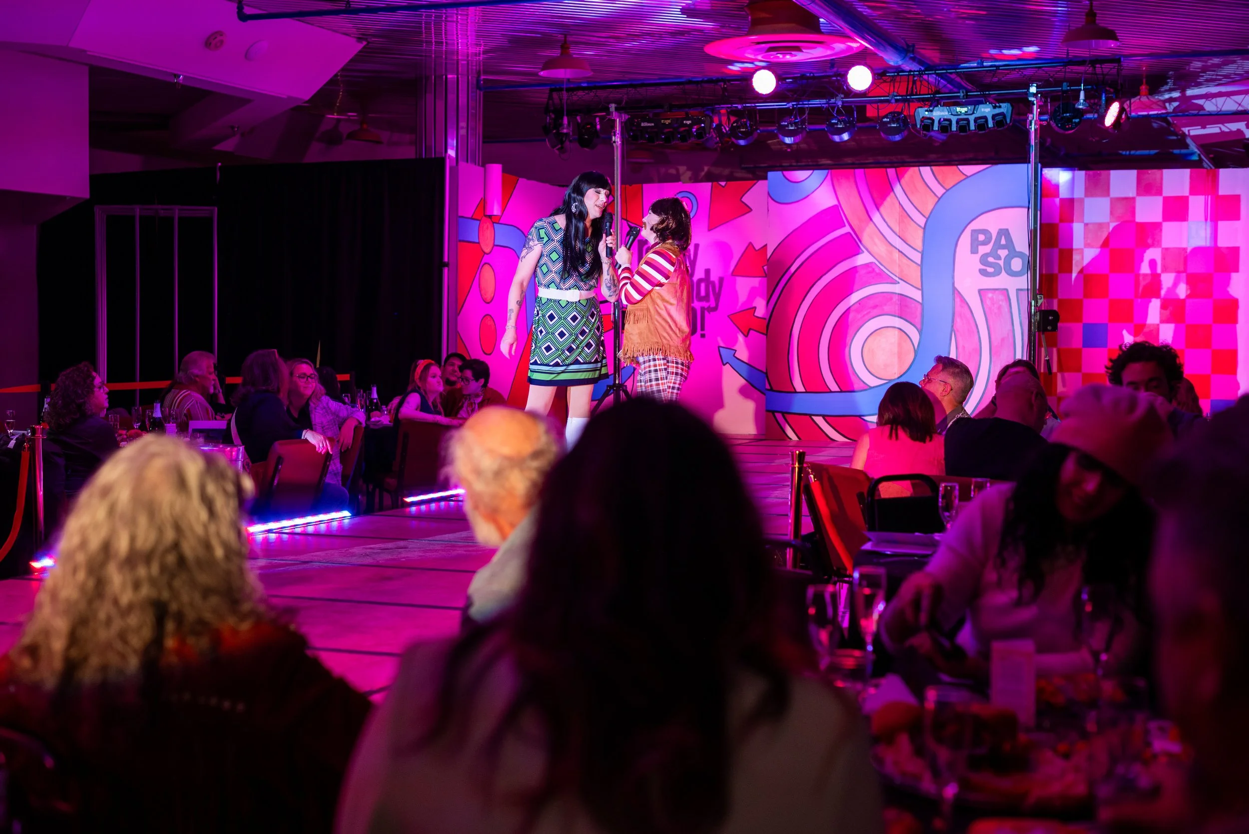 Stage performance at a lively event with two women singing into microphones, surrounded by seated audience.