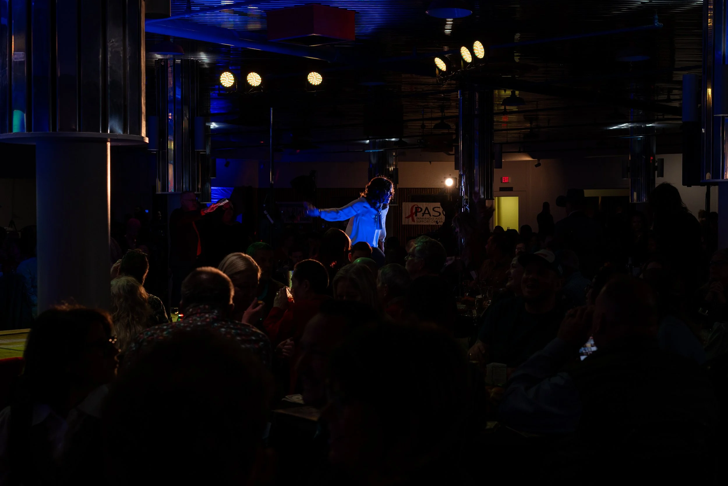 A dimly lit indoor event with a performer on stage illuminated by blue and white lights, surrounded by a seated audience.