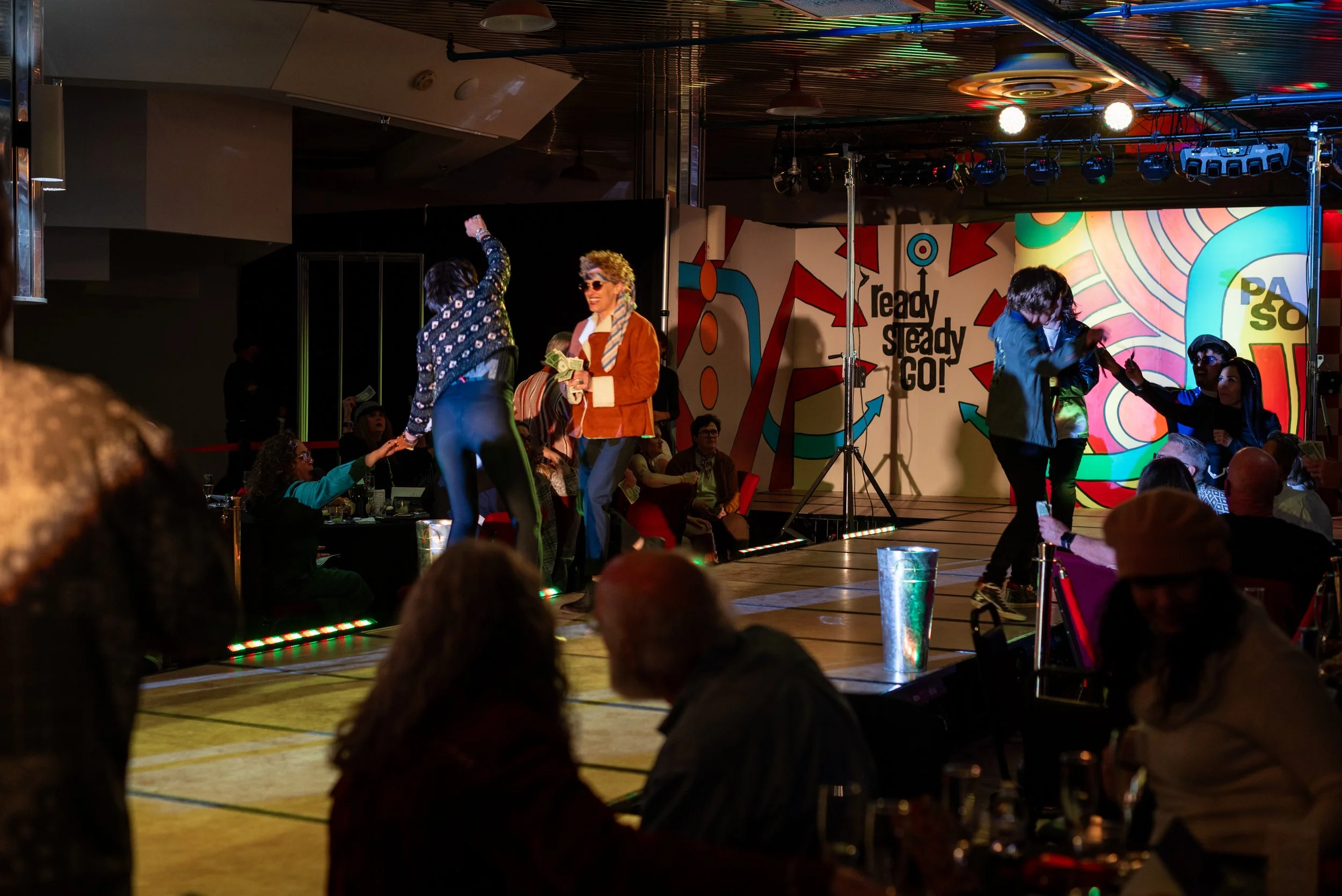 People dancing and enjoying a performance on a stage in an indoor entertainment venue with colorful lighting and a vibrant backdrop.