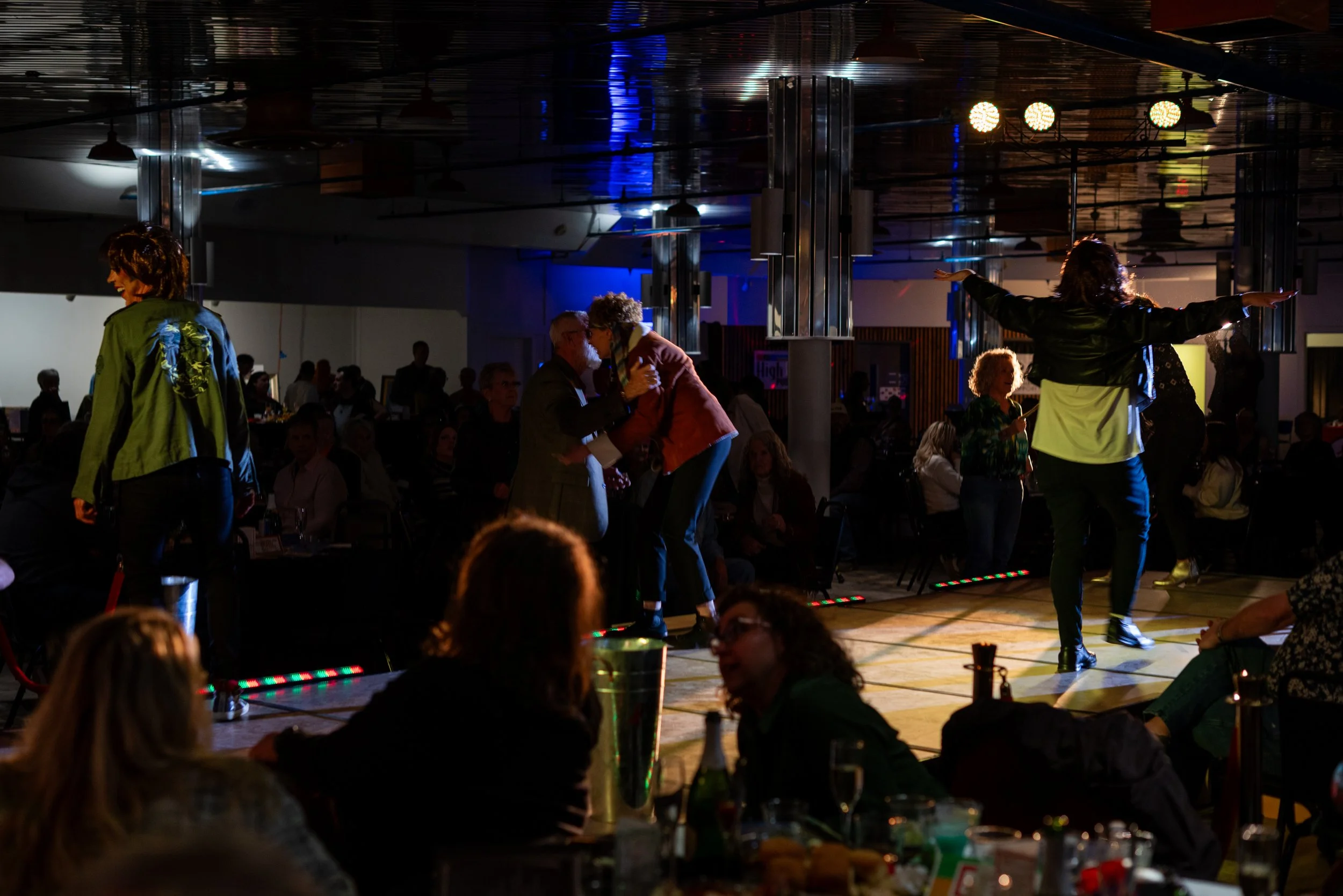 People dancing and socializing at a dimly lit indoor event, with some seated at tables in the foreground and others dancing on a lit dance floor in the background.