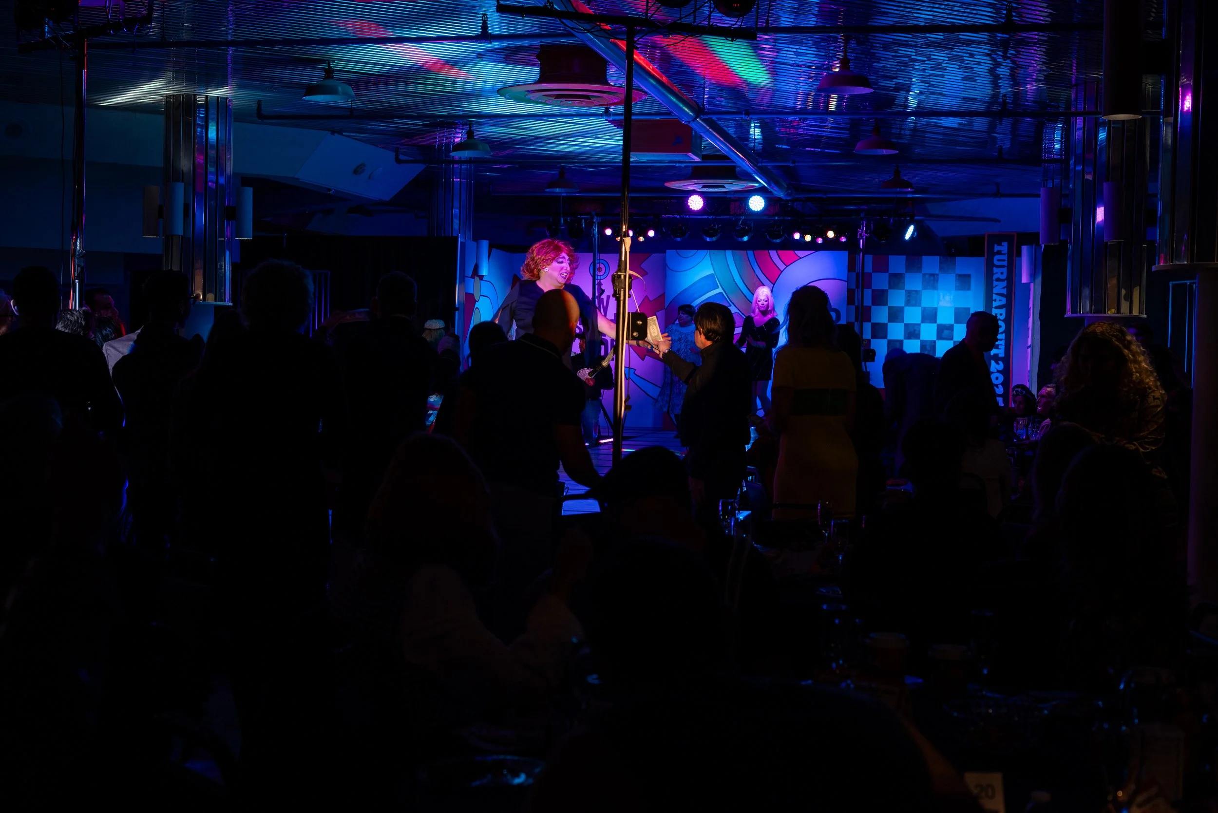 A dimly lit stage with performers and a woman receiving an award or certificate, with an audience watching, under colorful stage lighting.