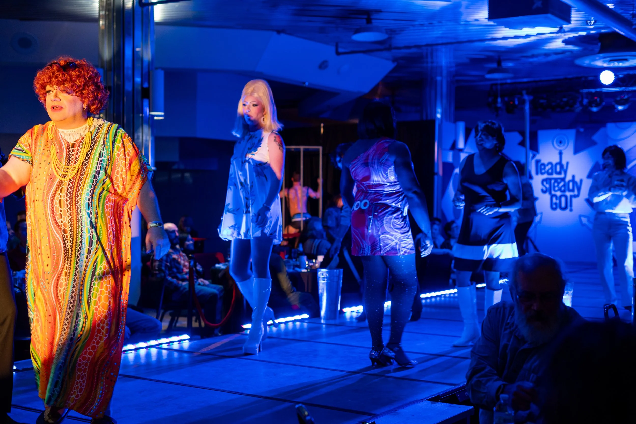 People on a stage performing or rehearsing in colorful outfits with a blue-lit background and a sign that reads 'Ready Steady Go!'