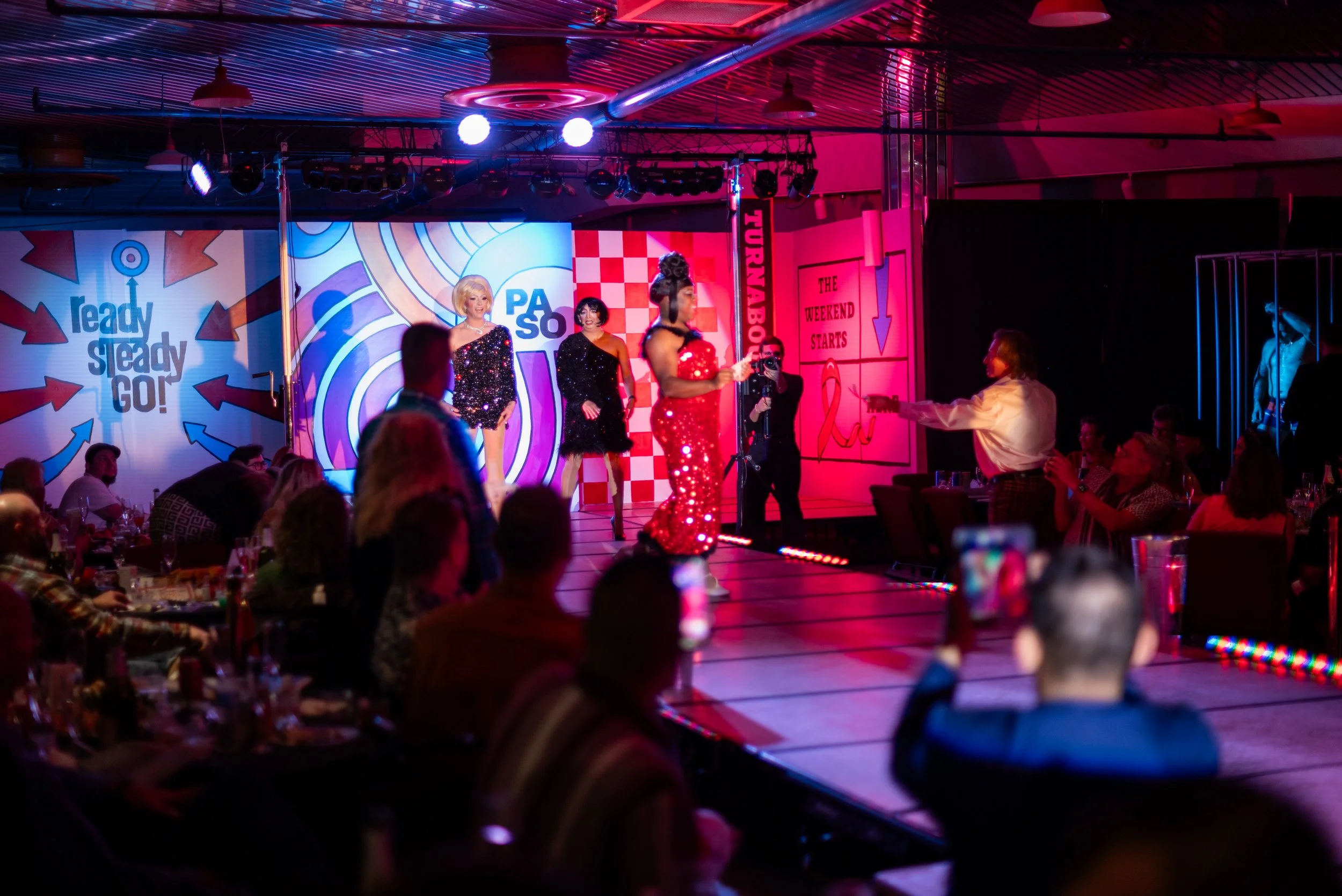 Drag performance on stage with three drag queens in sequined dresses, one in a red dress, one in a black dress, and one in a black and silver dress, with audience watching in a dimly lit venue with colorful signs and decorations.