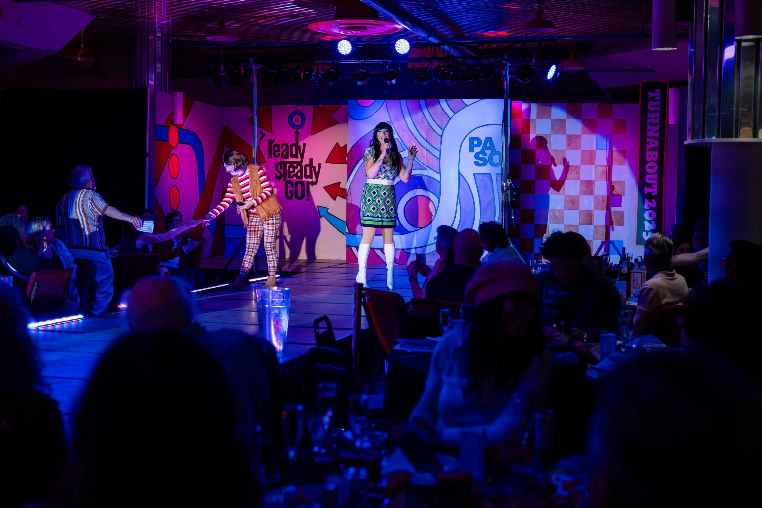 A performer on stage singing with a microphone in front of an audience in a dimly lit club. The background features colorful, abstract murals with phrases like "ready steady GO!" and "PASO". Some audience members are seated at tables, watching the pe