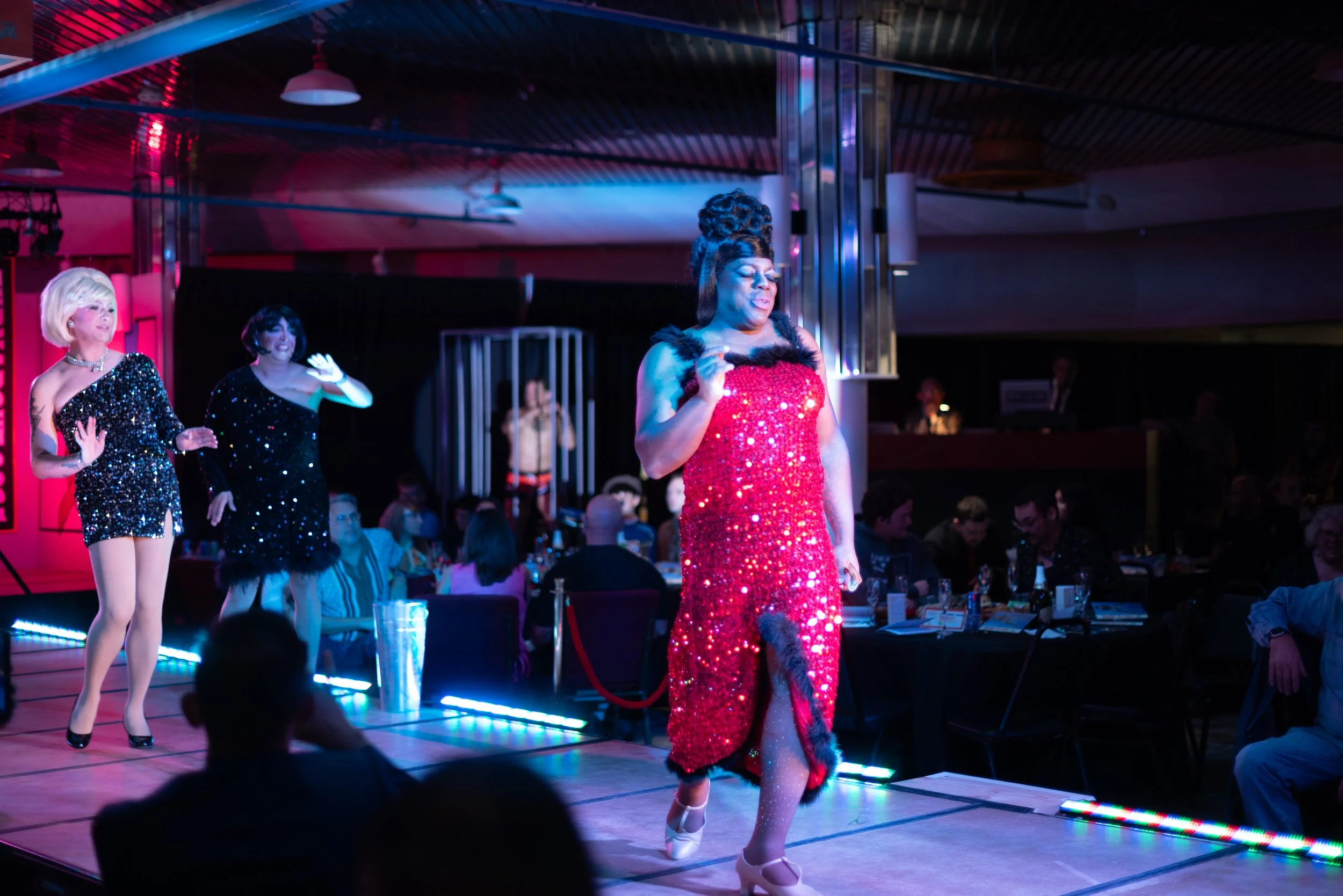 Three women performing on a stage at a drag show. The woman in the center wears a red, sequined dress with leopard print accents and high heels, while the women on the left wear black, glittery dresses with high heels. Audience members are seated at 