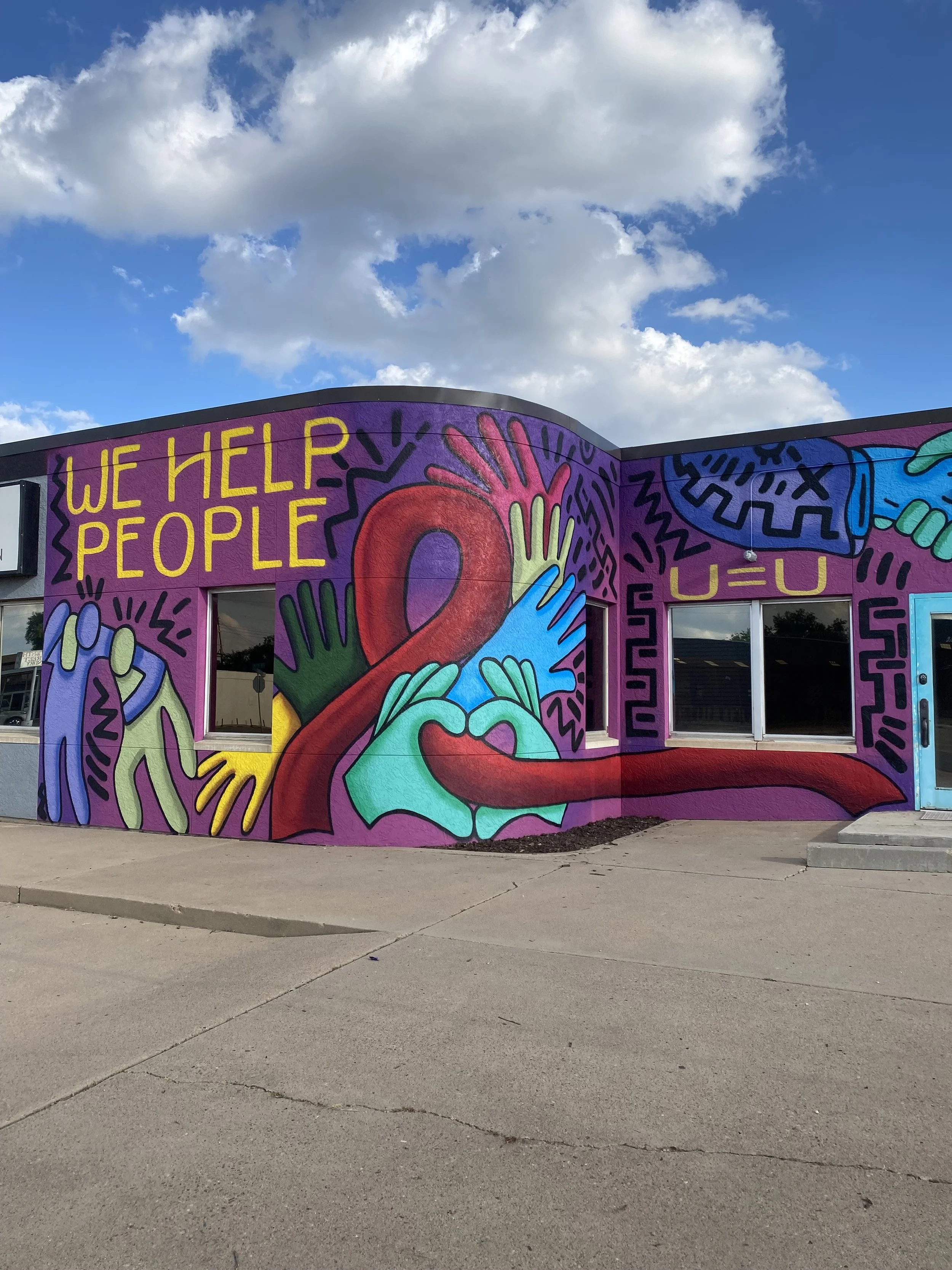 Colorful mural on a building with the words "We Help People." The mural features abstract human figures, hands, and heart shapes with vibrant colors like purple, blue, green, red, and yellow, set against a bright sky with scattered clouds.