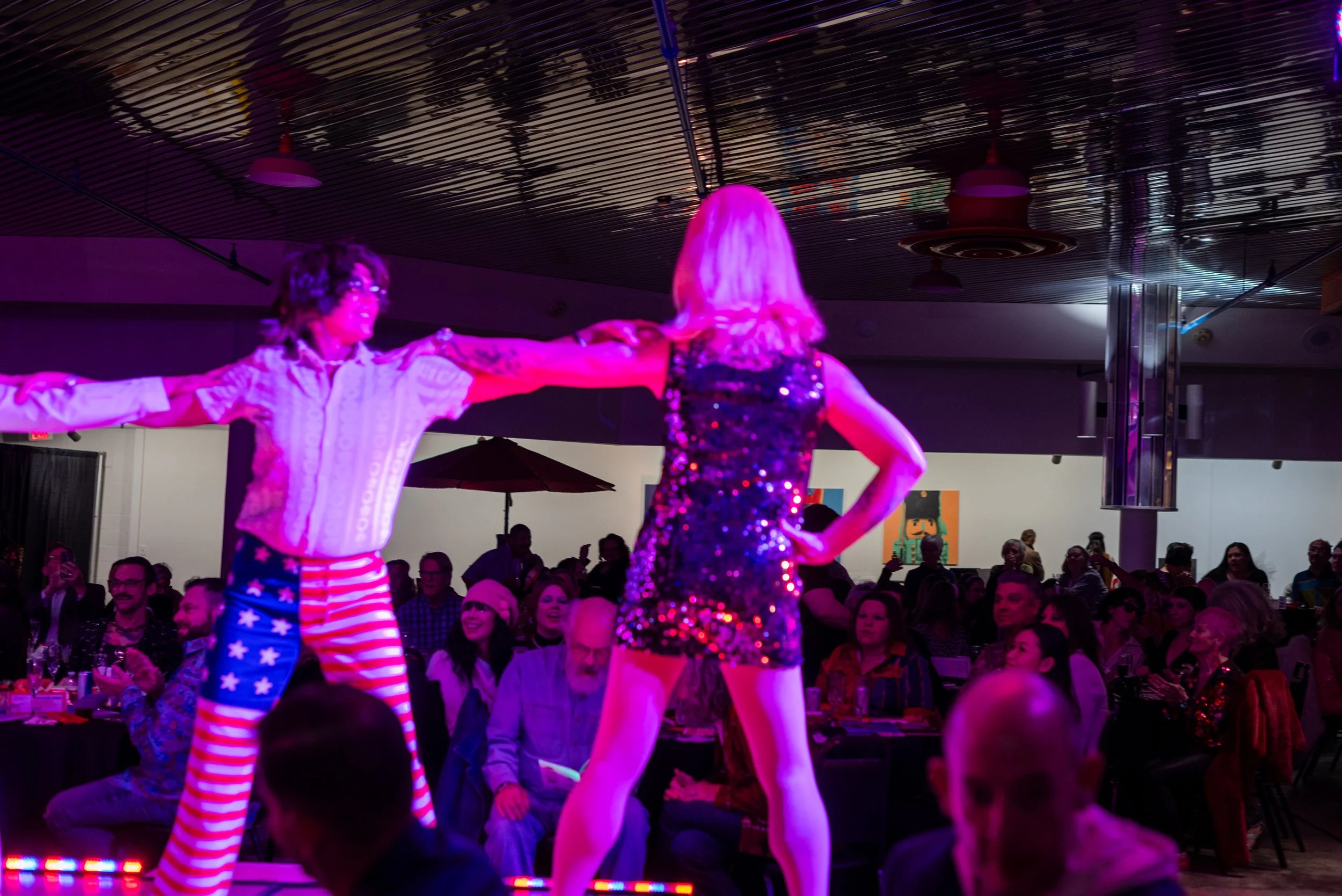 Two performers dancing on stage with audience watching in a dimly lit setting. One performer wears patriotic-themed pants and a white shirt, the other wears a sparkling black dress.