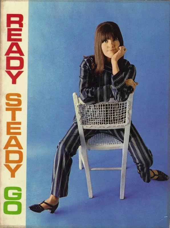 A woman seated on a white chair against a blue background, wearing striped pajamas, with her right hand resting on her chin and looking at the camera. The title 'Ready to Stay' is vertically written in multicolored letters on the left side.