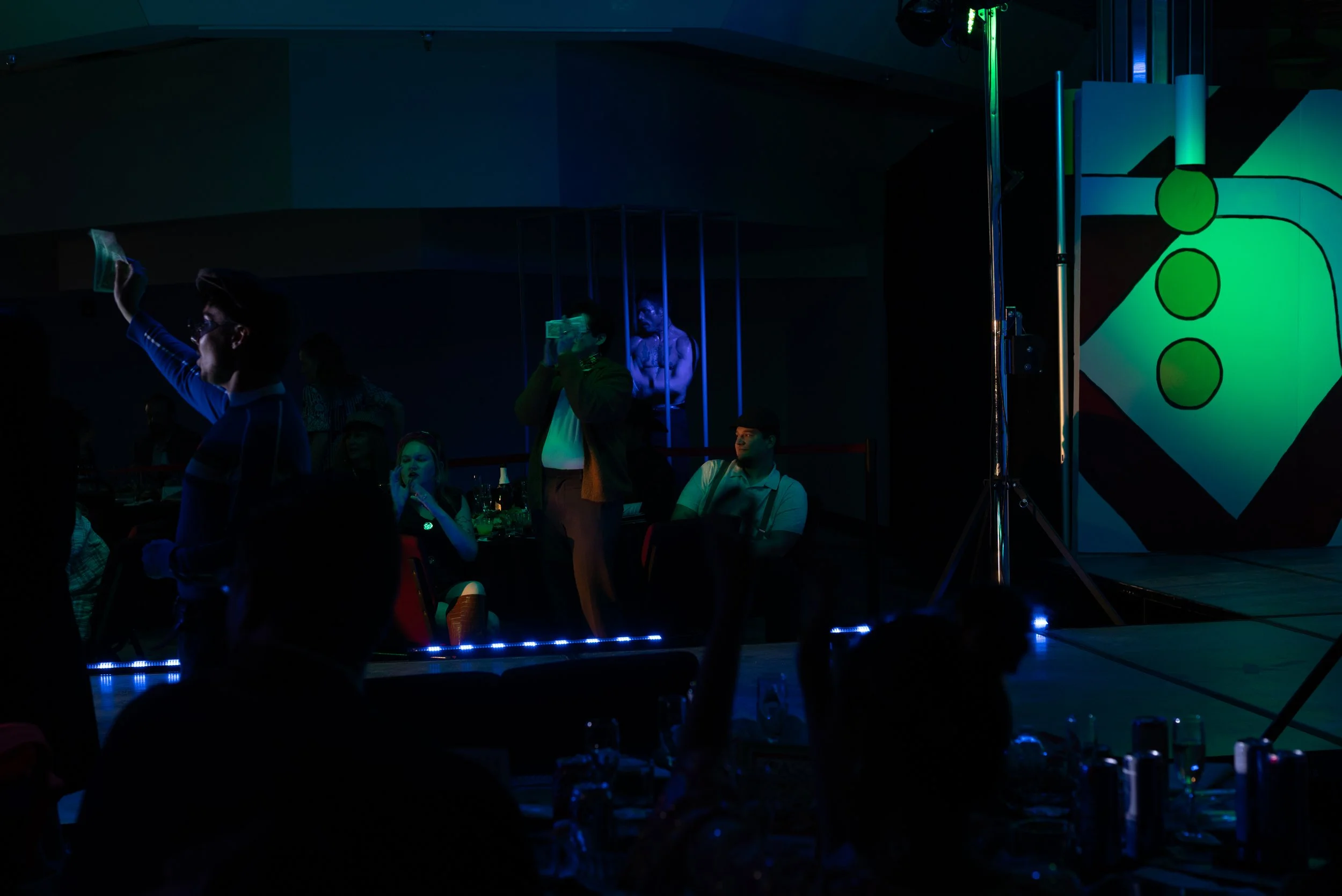 Scene from a dark, lively event with people sitting at tables, some standing, and a performer waving a towel on stage illuminated by blue and green lights, with a large art piece featuring a domino and green circles in the background.