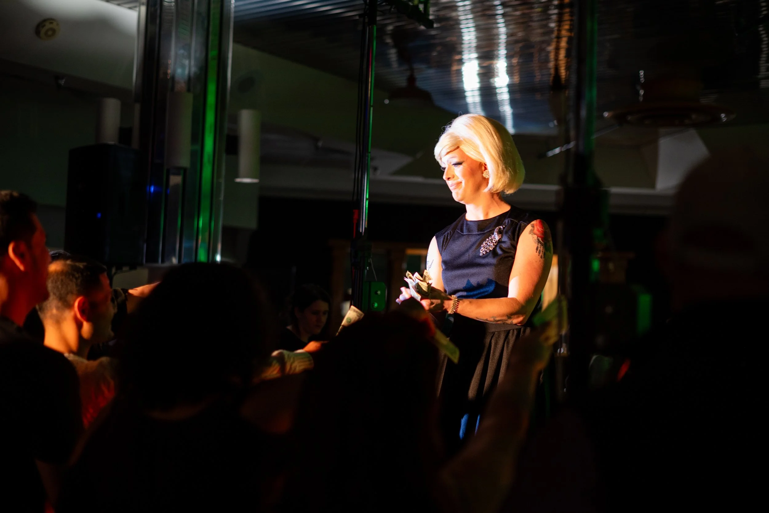 A woman with blonde hair, wearing a black dress with tattoos on her arms, stands on stage with a microphone in her hand, illuminated by stage lights, while an audience looks on in a dark room.