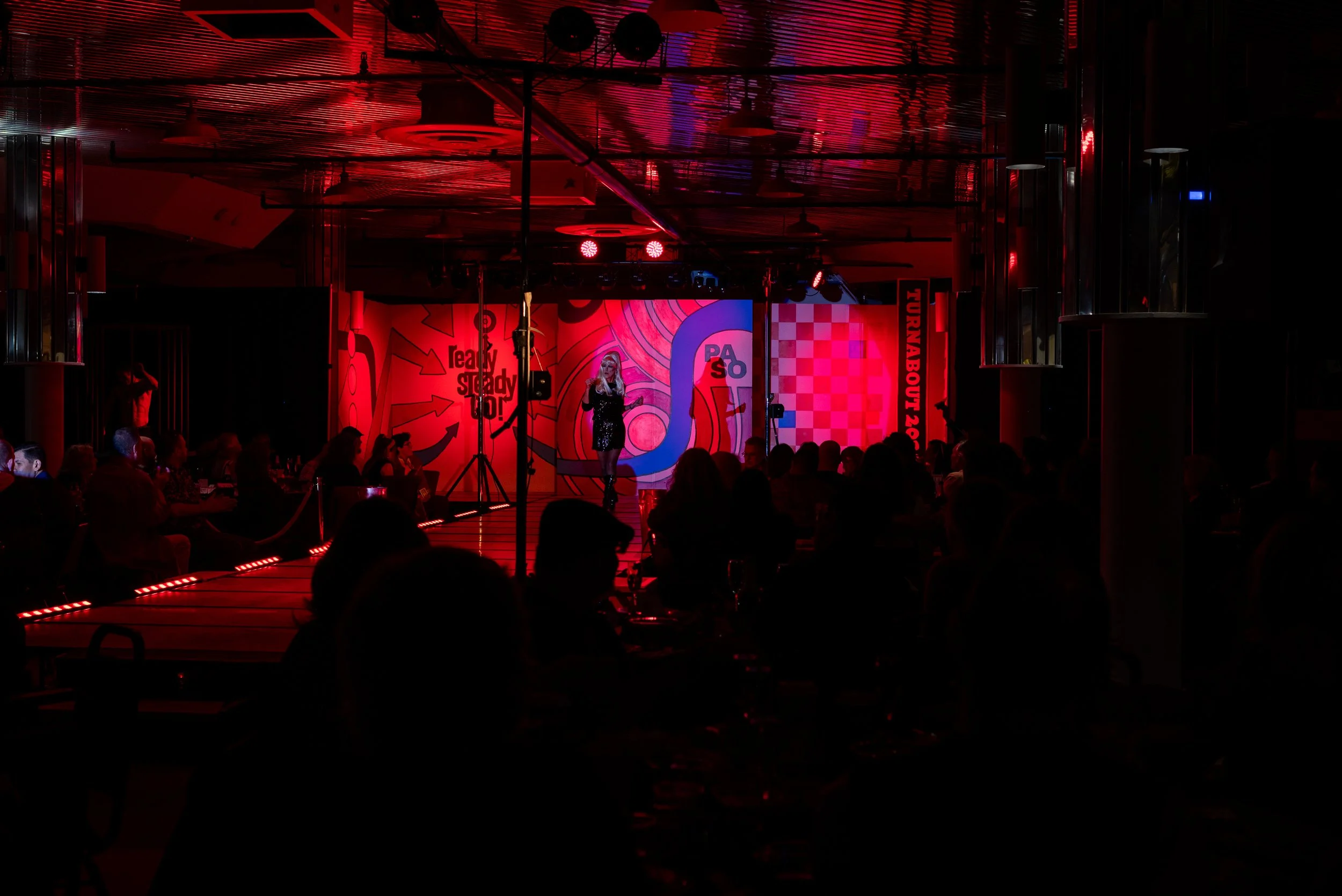 A woman performing on a stage with red lighting, in front of an audience seated at tables in a dark venue with colorful backdrops.