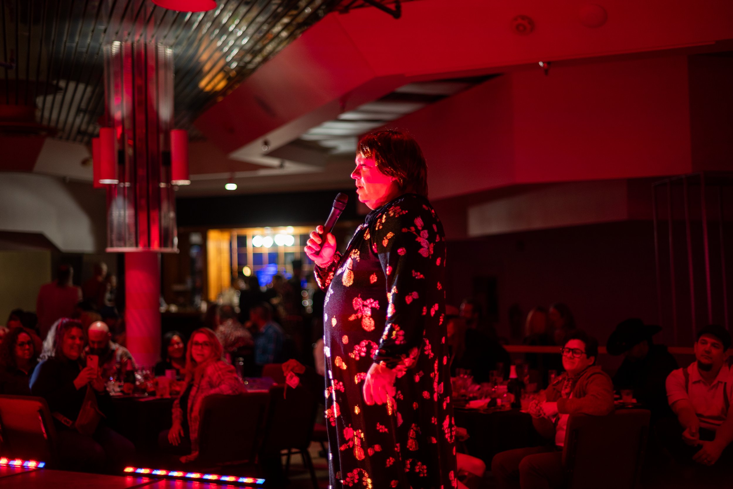 A woman on stage holding a microphone during a performance or comedy show, with an audience seated at tables watching her.
