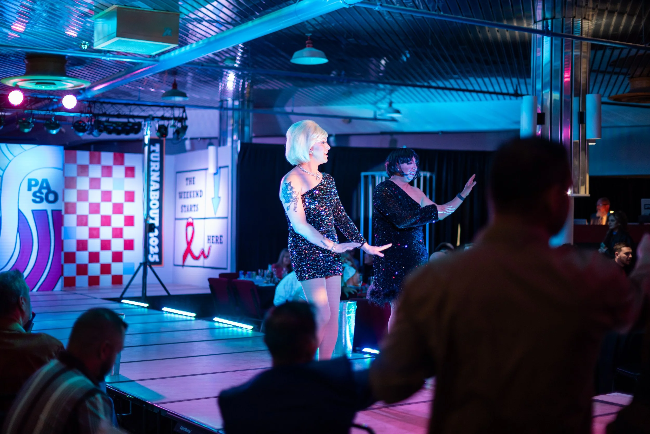 Two performers dancing on a stage with colorful lights at a fundraiser event, with audience members watching.