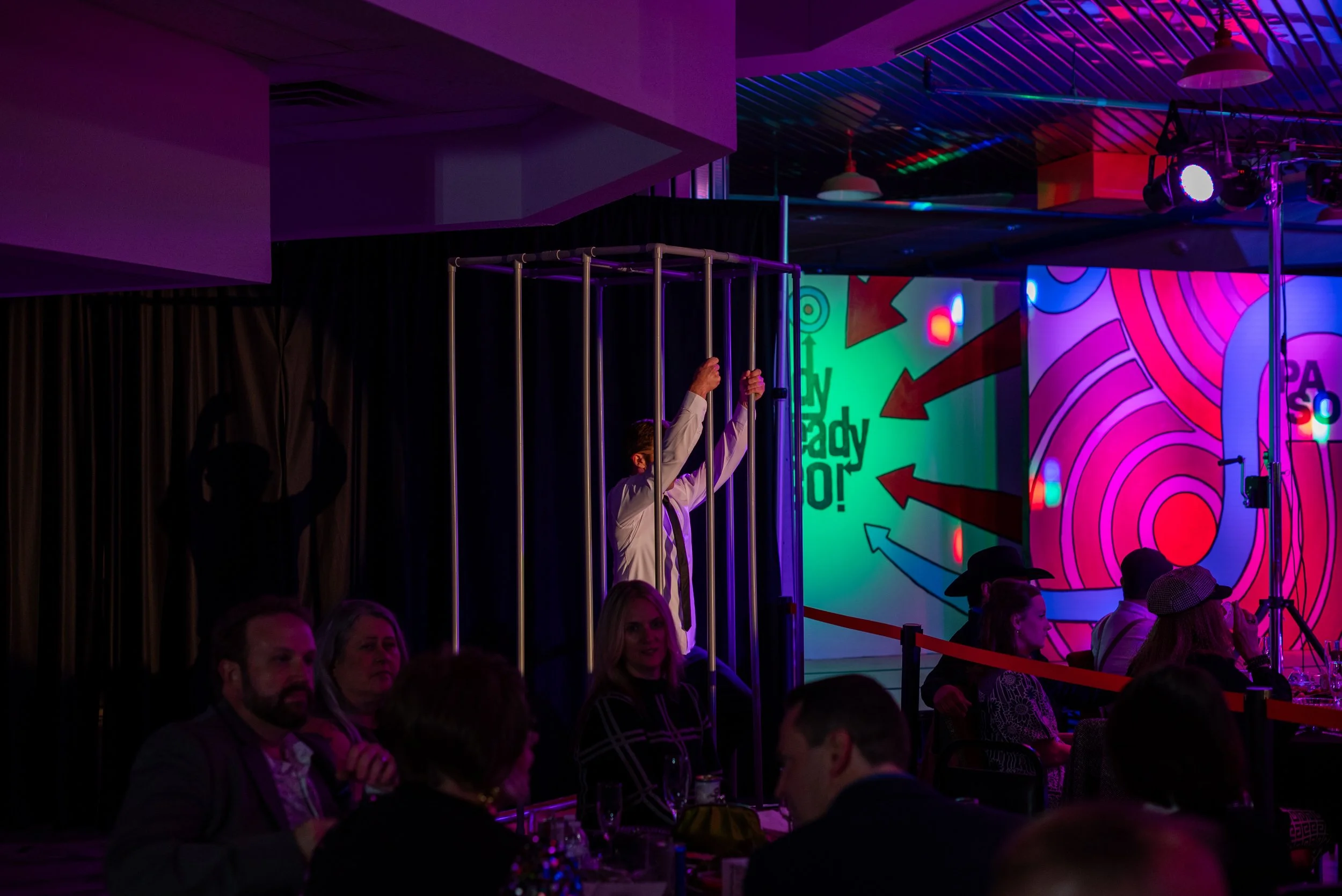 A man standing inside a metal cage at an event with colorful abstract wall art and audience members seated at tables.