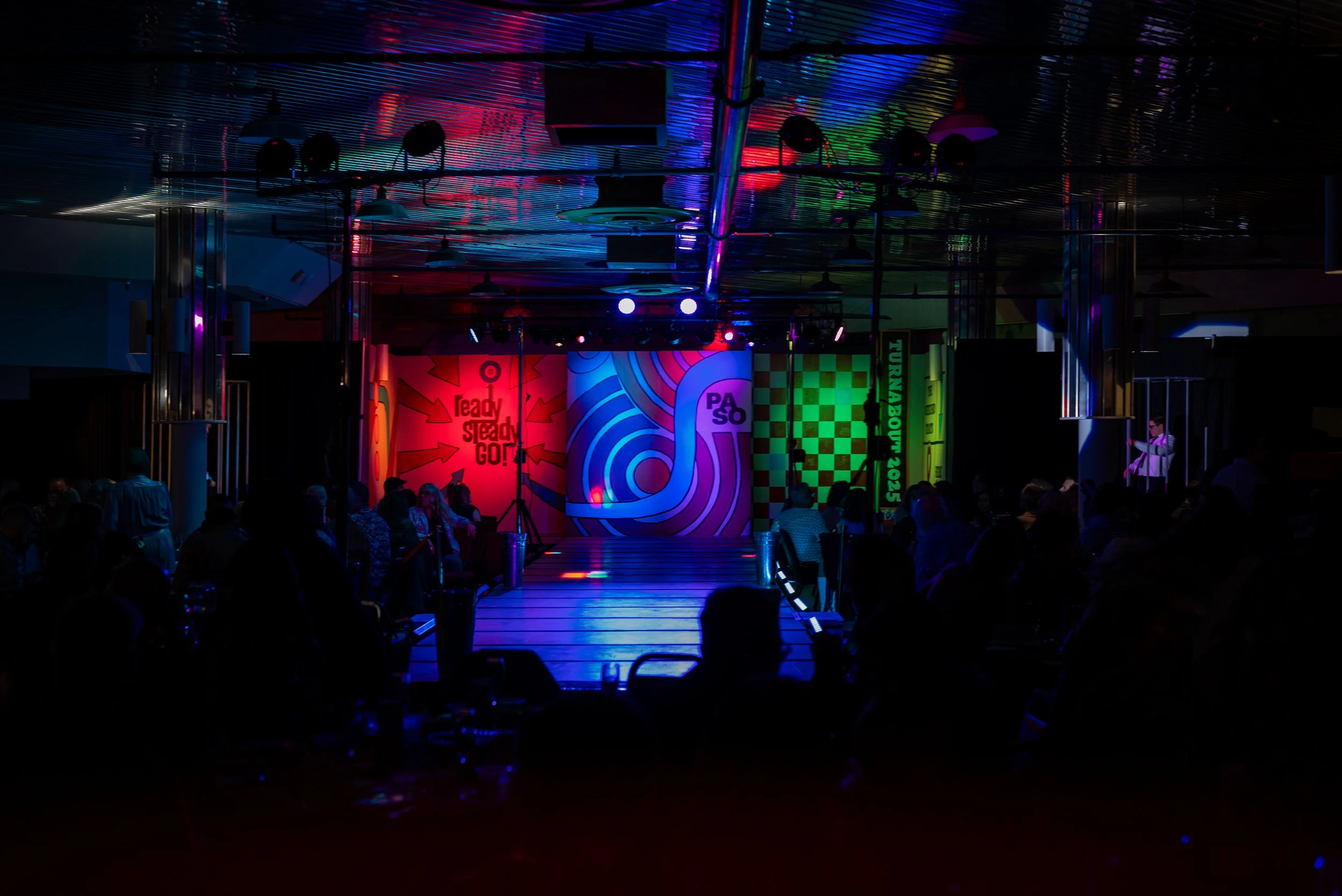 Indoor event space with a stage and colorful, abstract backdrops, including the texts 'Ready Steady Go!' and 'PASO,' illuminated by vibrant lighting, with an audience seated in the dark.