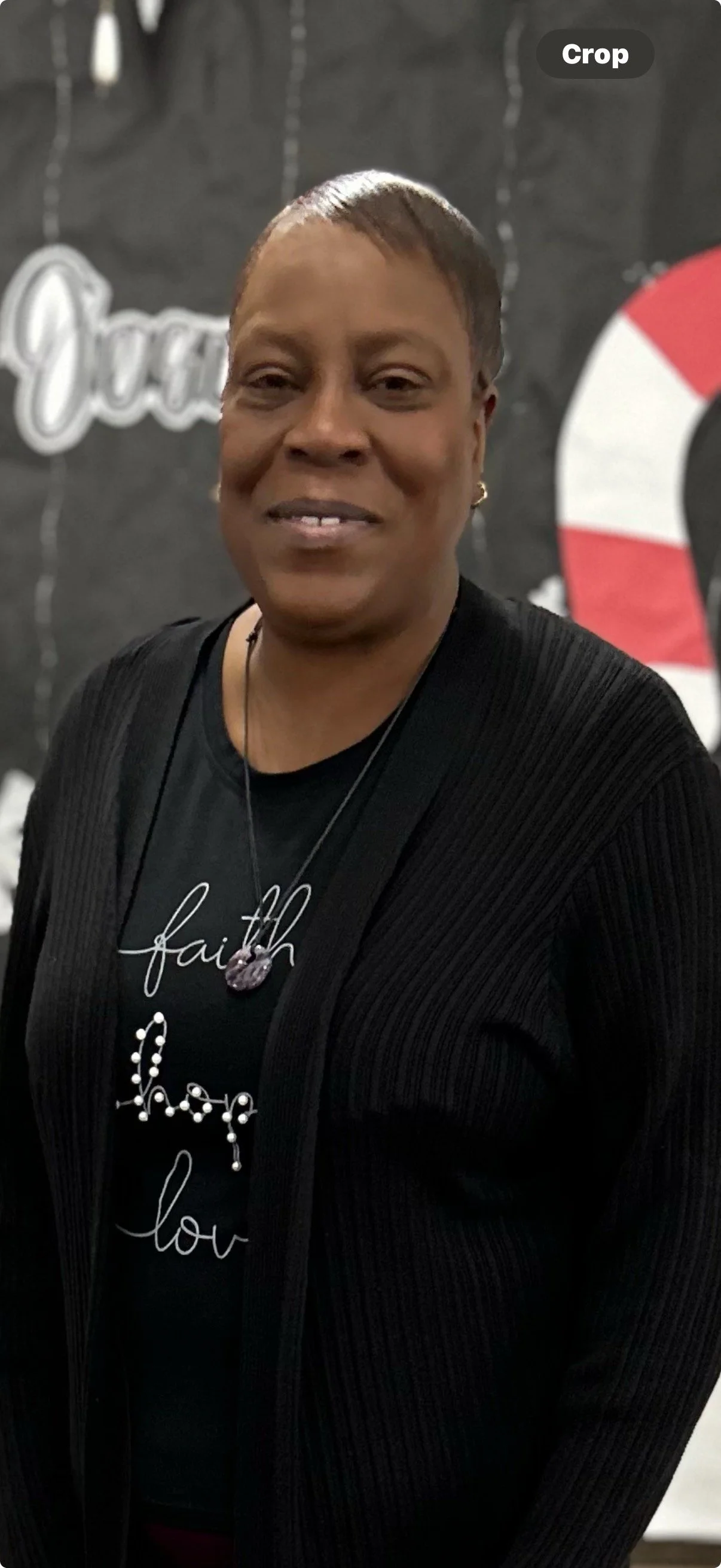 A woman with short hair wearing a black cardigan and a black t-shirt with white cursive text and decorative design, standing in front of a black background with Christmas-themed decorations.