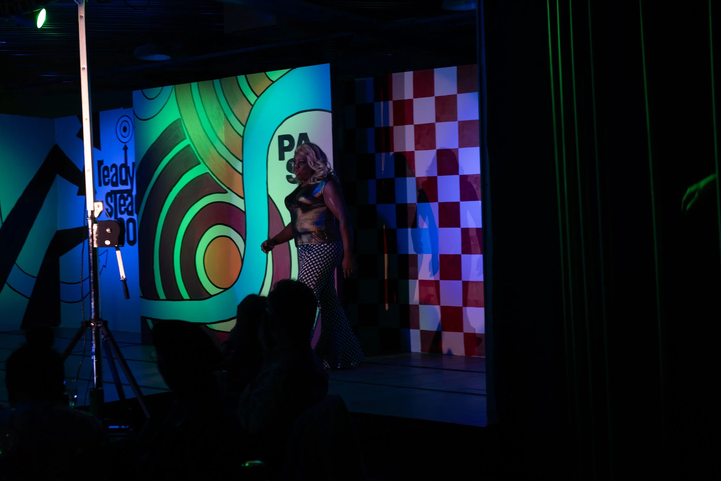 Performer on stage with colorful abstract background, audience seated in front, dark lighting with stage spotlights.
