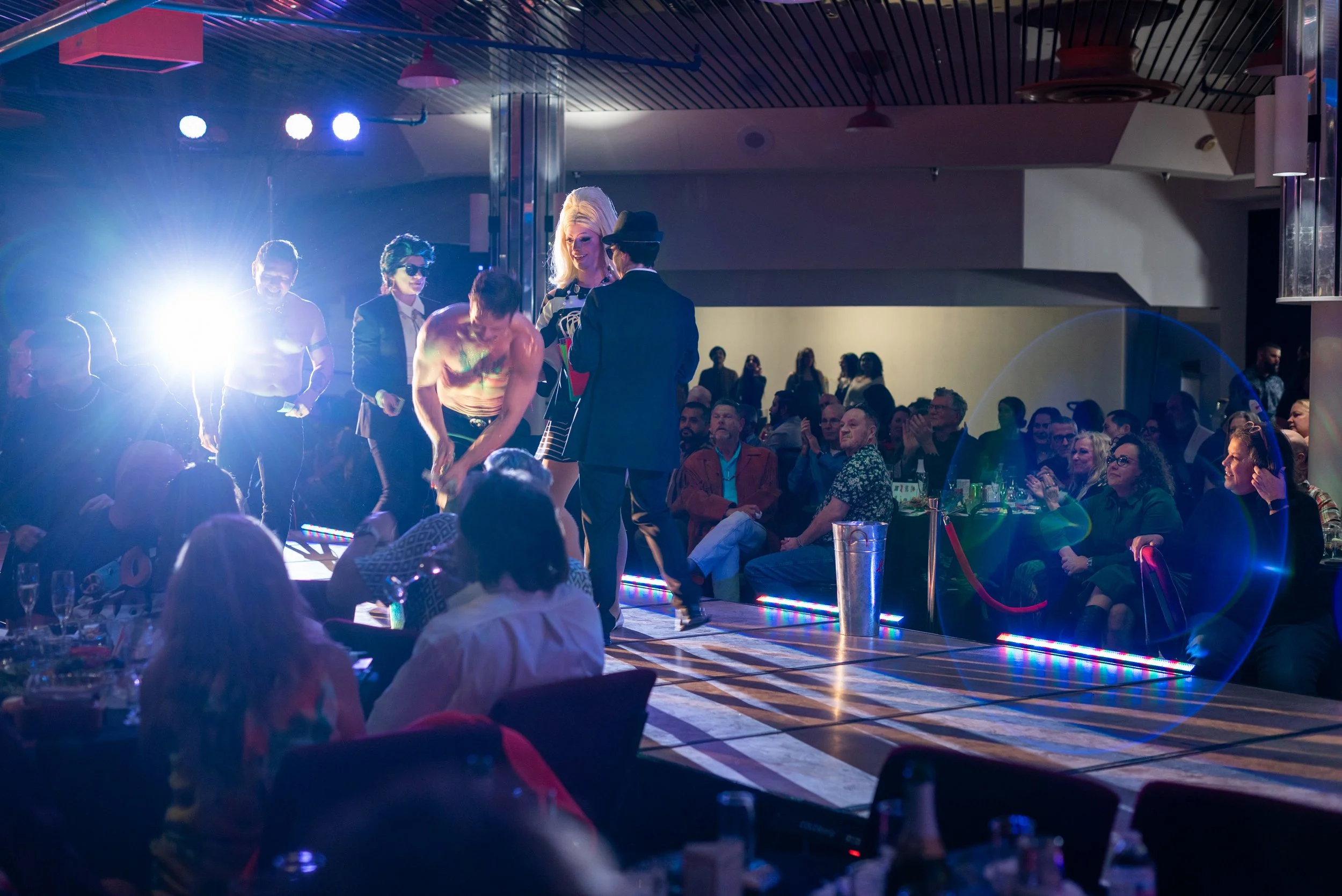 People performing on a stage with an audience watching in a dimly lit theater with colorful lights and concert equipment.