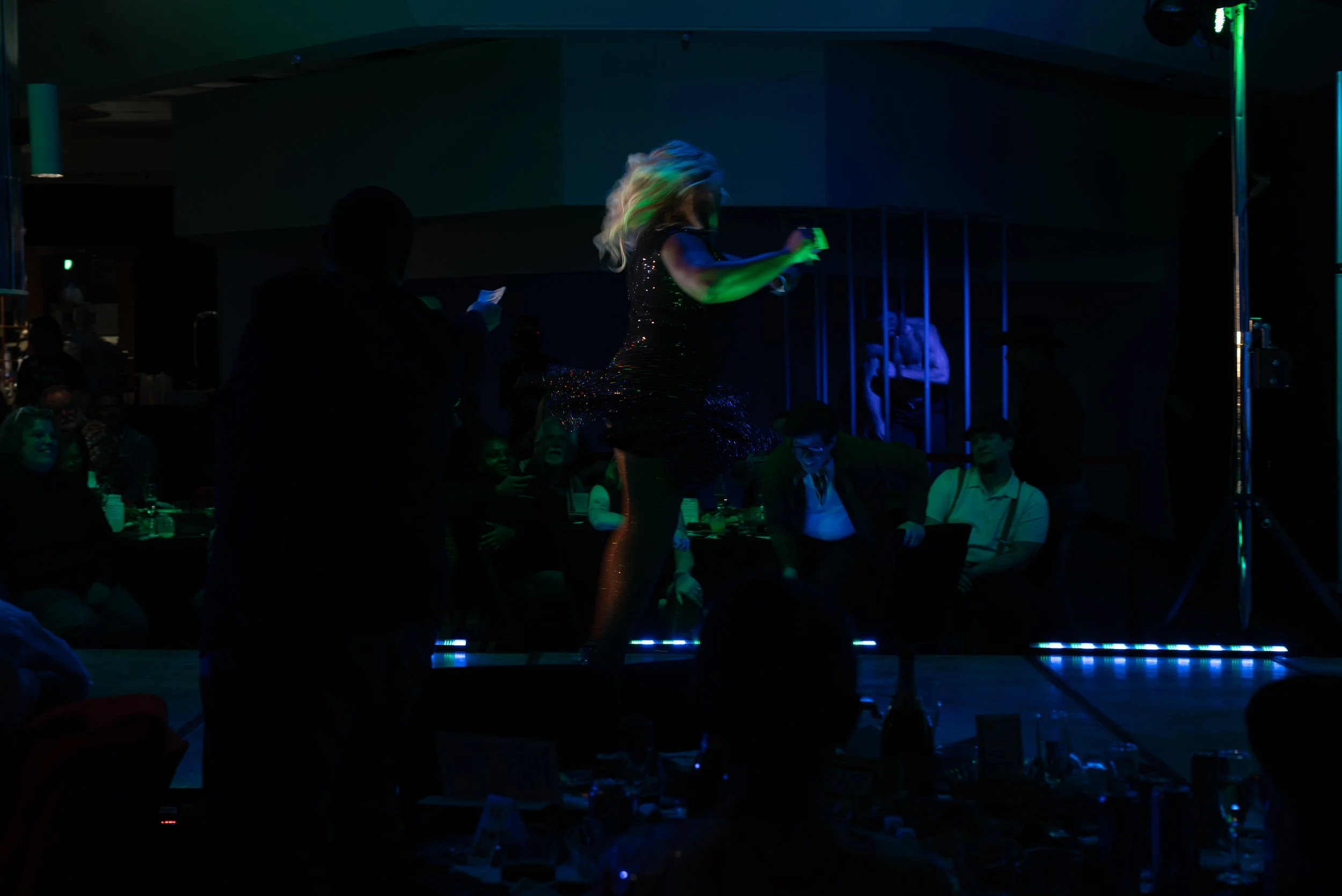 A woman performing a dance on a stage in front of an audience at a dark, colorful event.