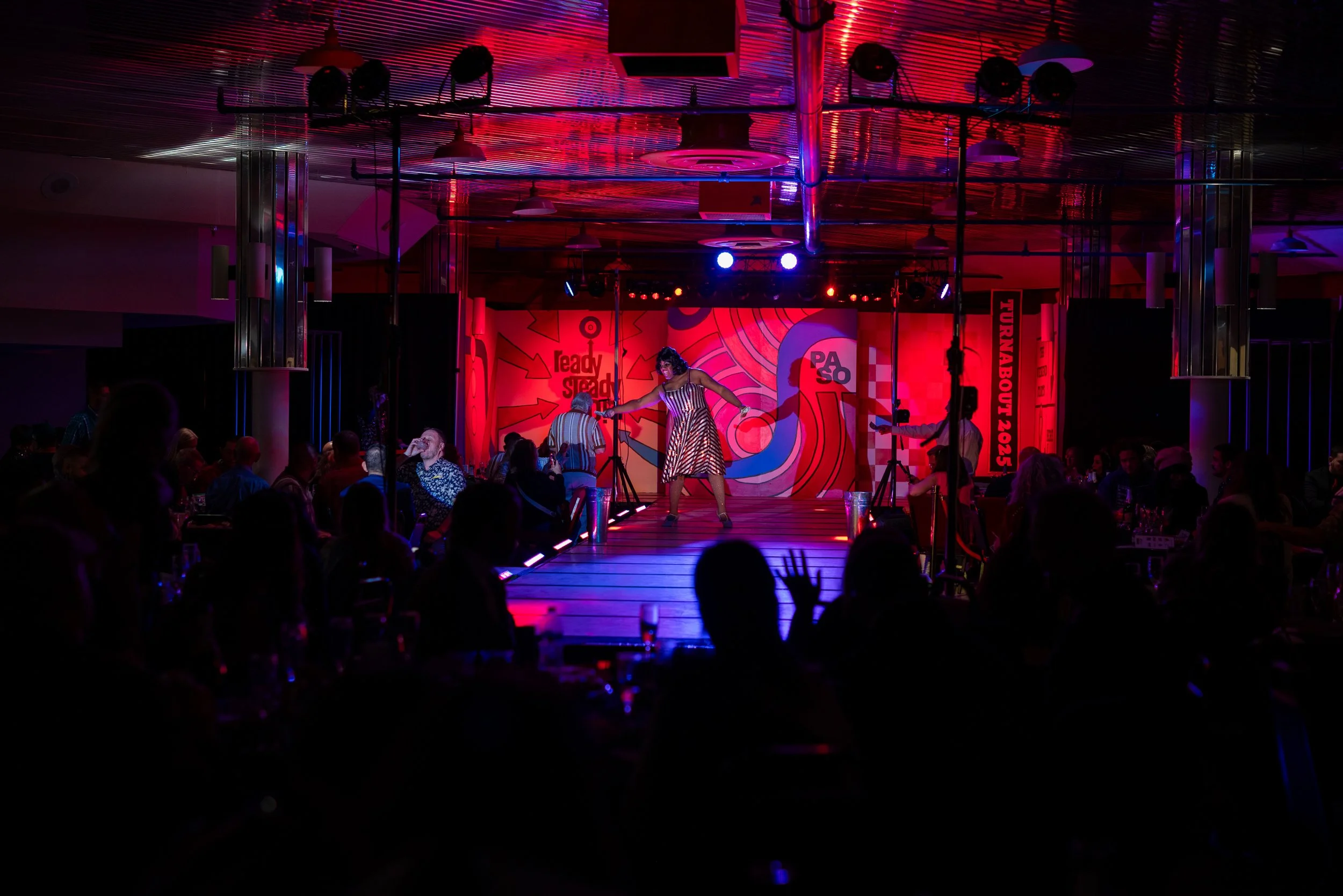 Performer on a stage with colorful, abstract backdrop, singing and dancing in a vibrant indoor event with seated audience, illuminated by stage lights, at night.